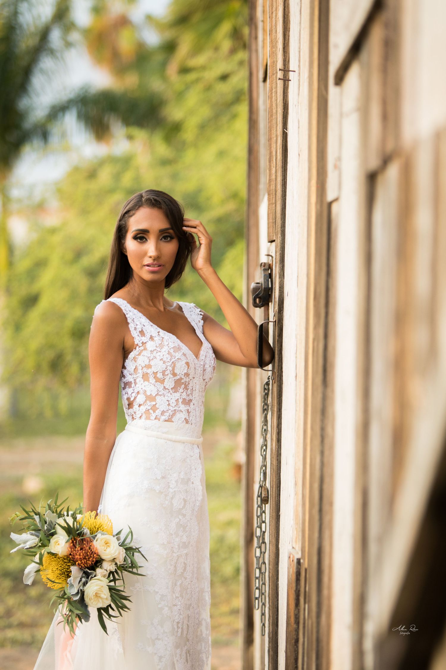 Bride in lace wedding dress holding bouquet leaning against rustic wooden door outdoors.