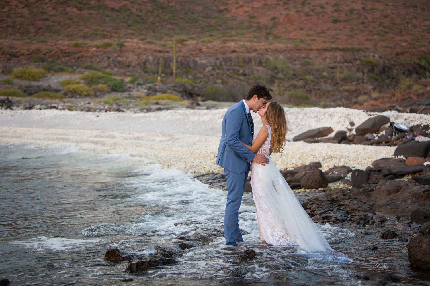 Bride and groom share romantic kiss at rocky shoreline with gentle ocean waves washing around their feet.