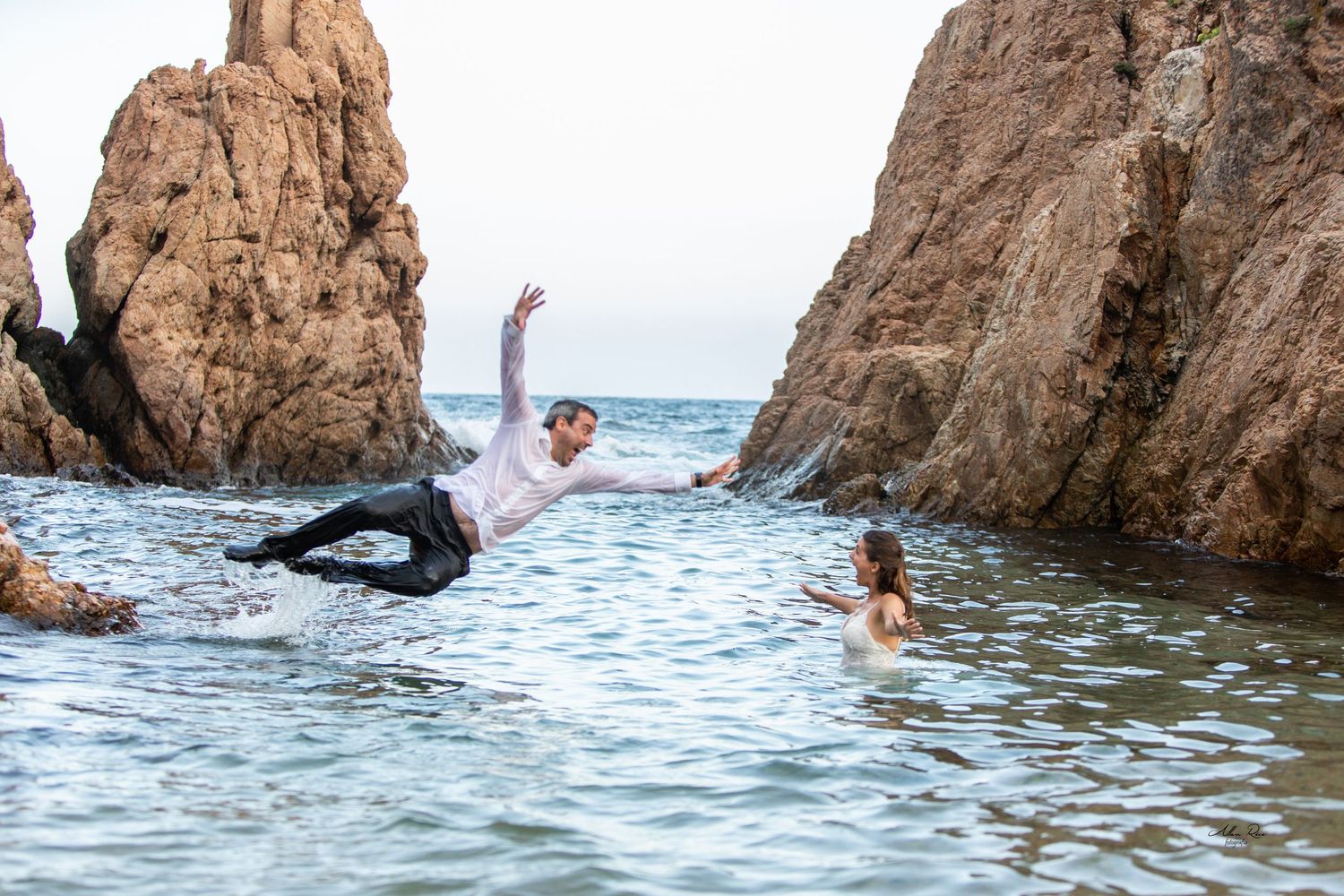 Groom playfully falls into ocean water as bride watches between scenic coastal rocks.