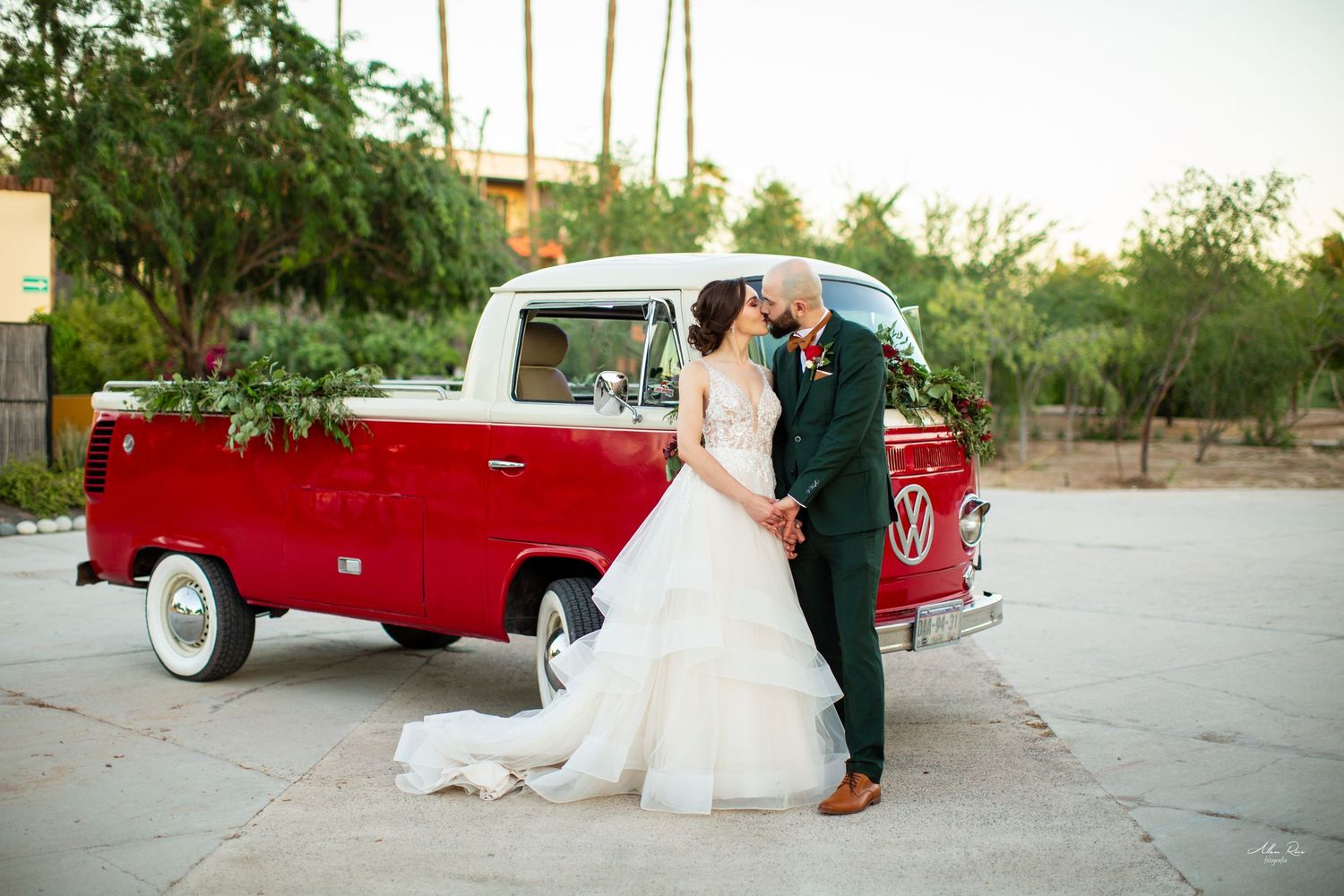 Bride and groom kissing in front of vintage red Volkswagen bus at outdoor wedding venue.