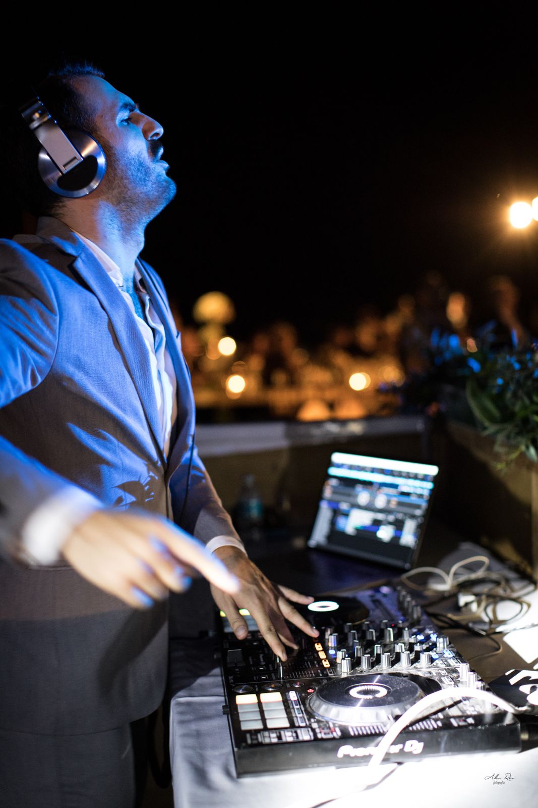 Professional DJ in gray suit with headphones mixing music at turntable during outdoor nighttime wedding reception.