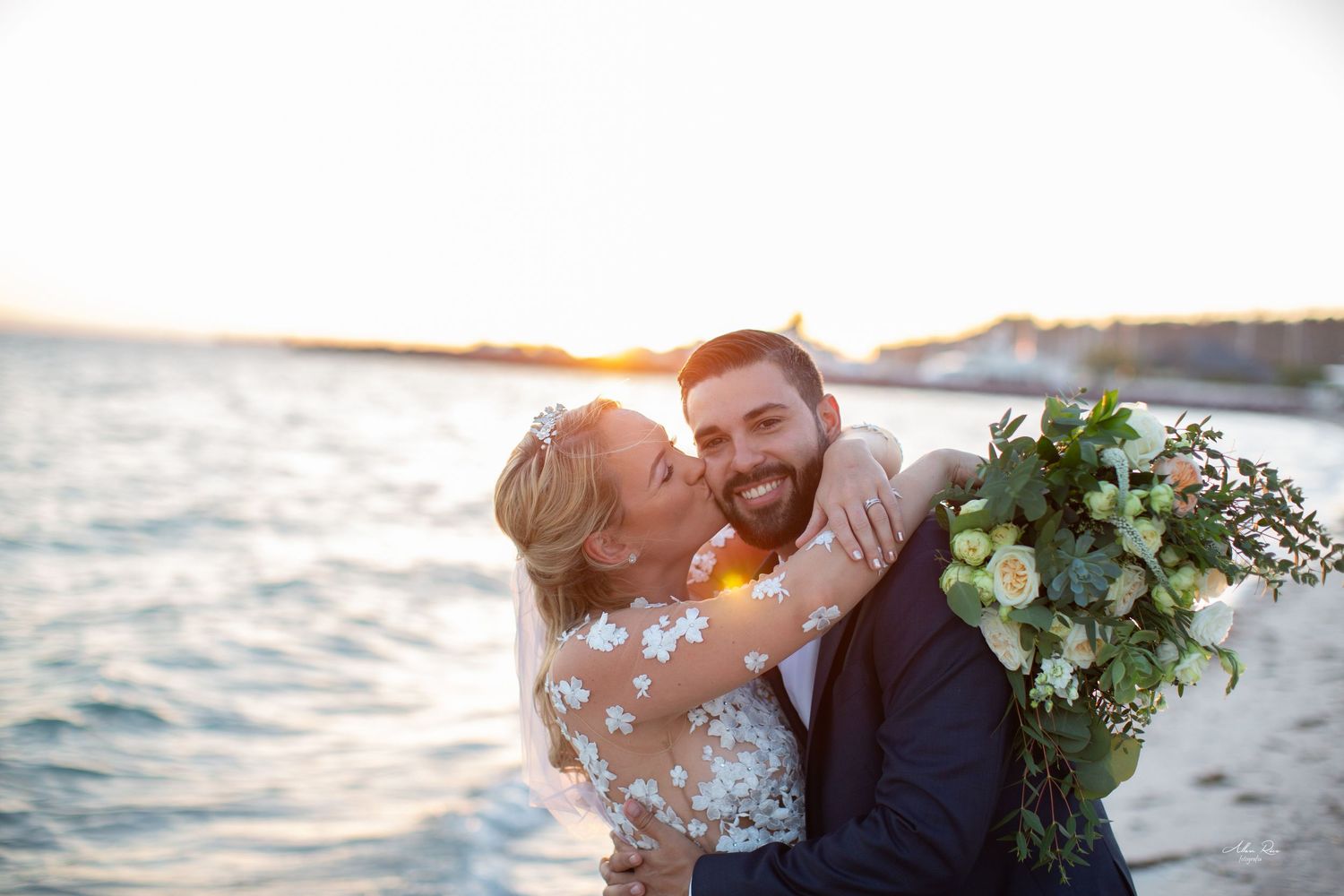 Joyful bride kissing groom on cheek at golden hour sunset with ocean backdrop and bouquet.