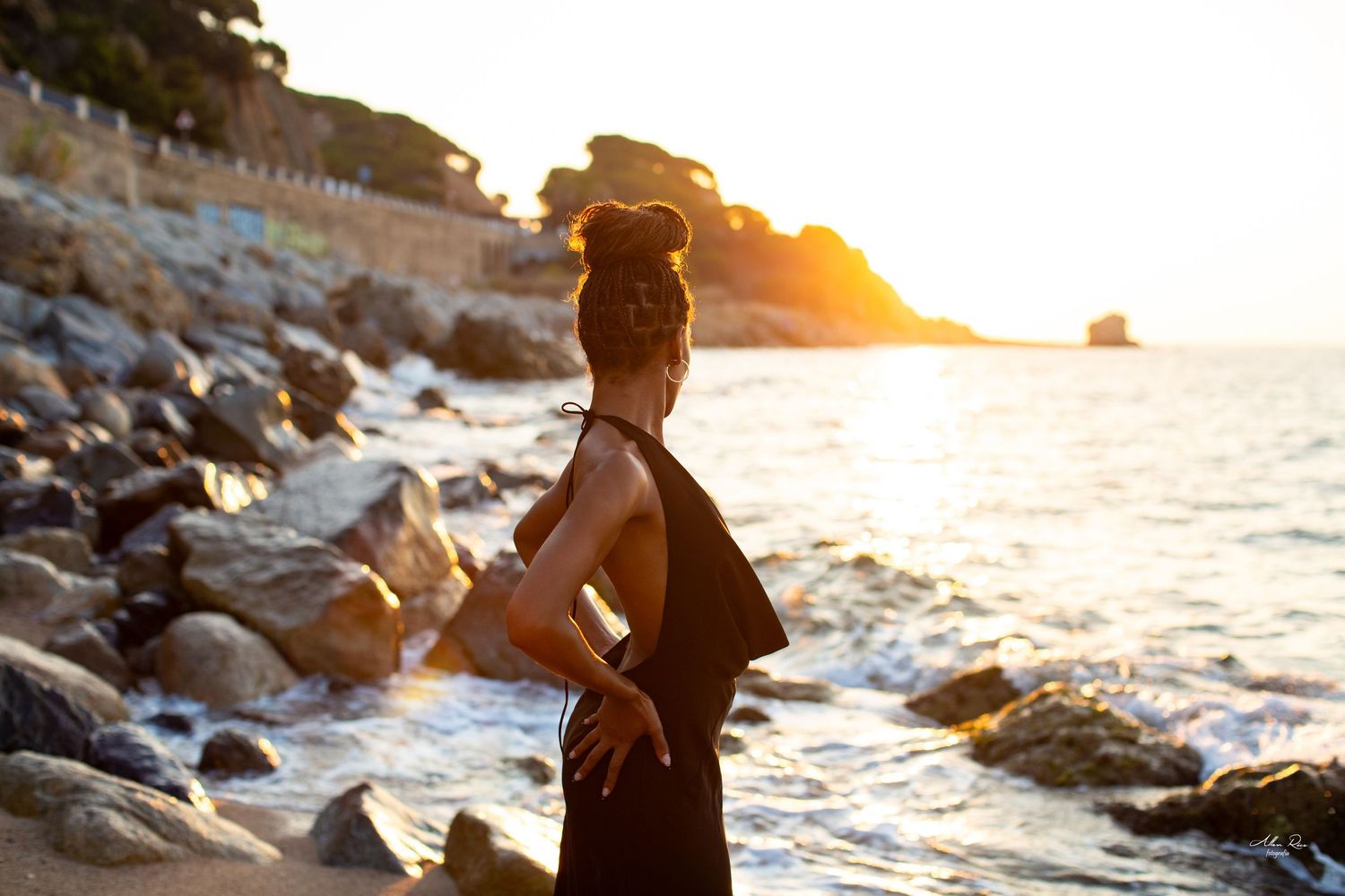 Woman in black dress standing on rocky beach at golden sunset with ocean waves and coastal cliffs in background.