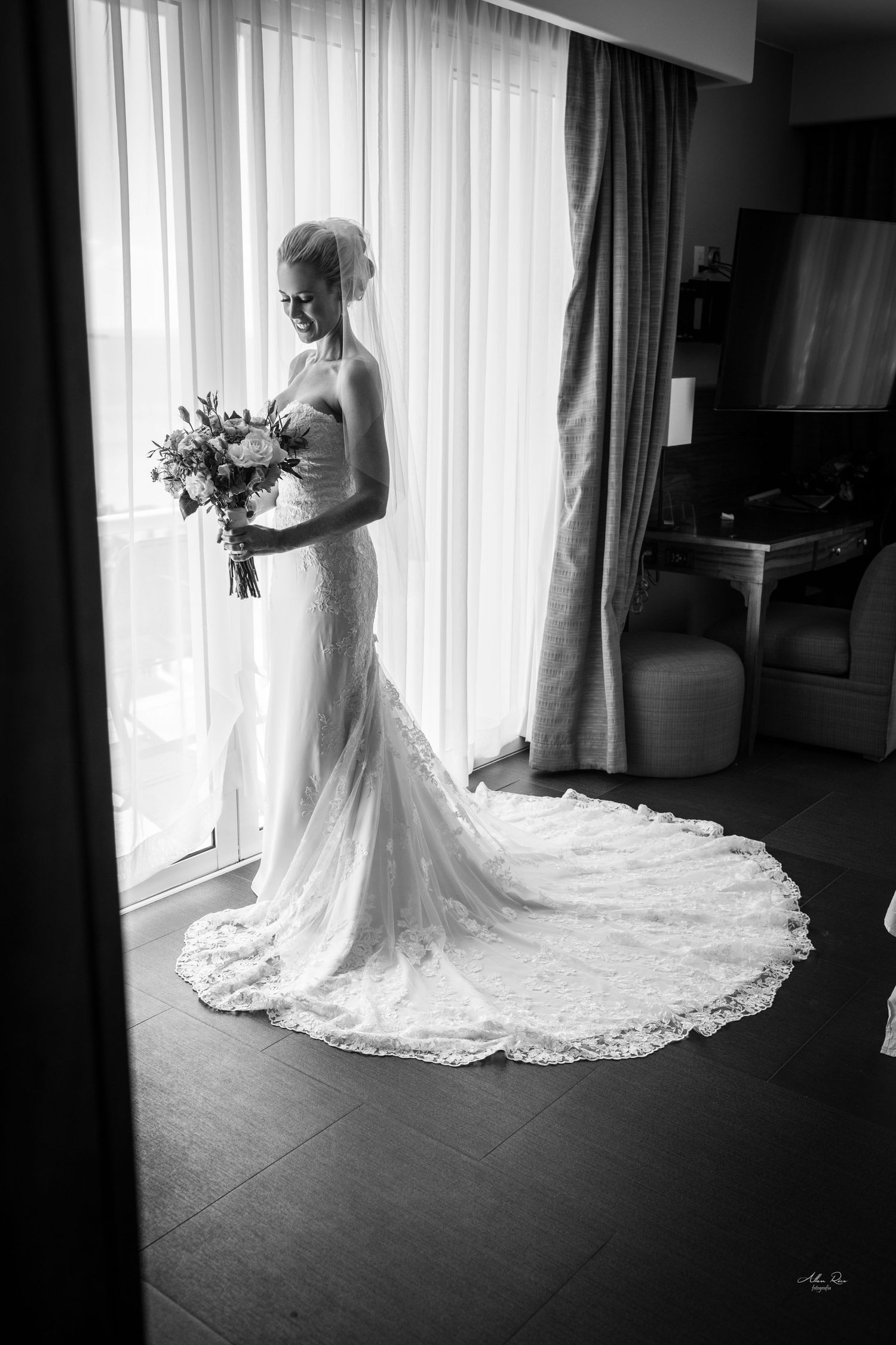 Bride in a lace wedding gown holding a bouquet, standing by a window in black and white.