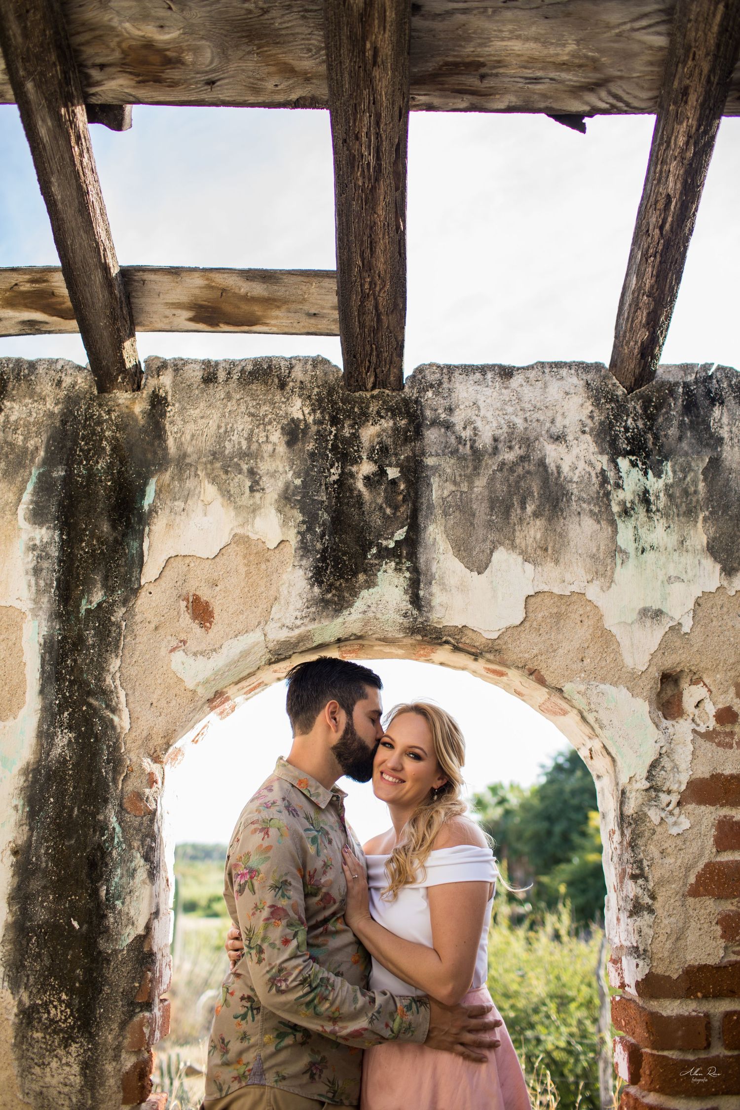 Couple embracing romantically inside rustic stone archway ruins with wooden beam ceiling.