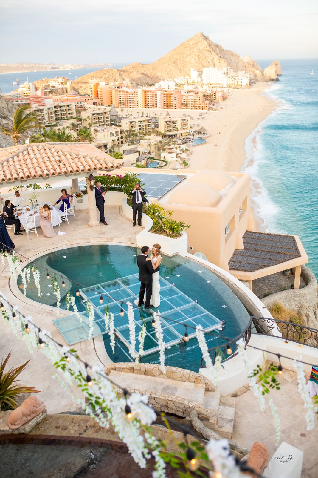 Couple sharing first dance on glass floor over pool with stunning Cabo San Lucas coastline.
