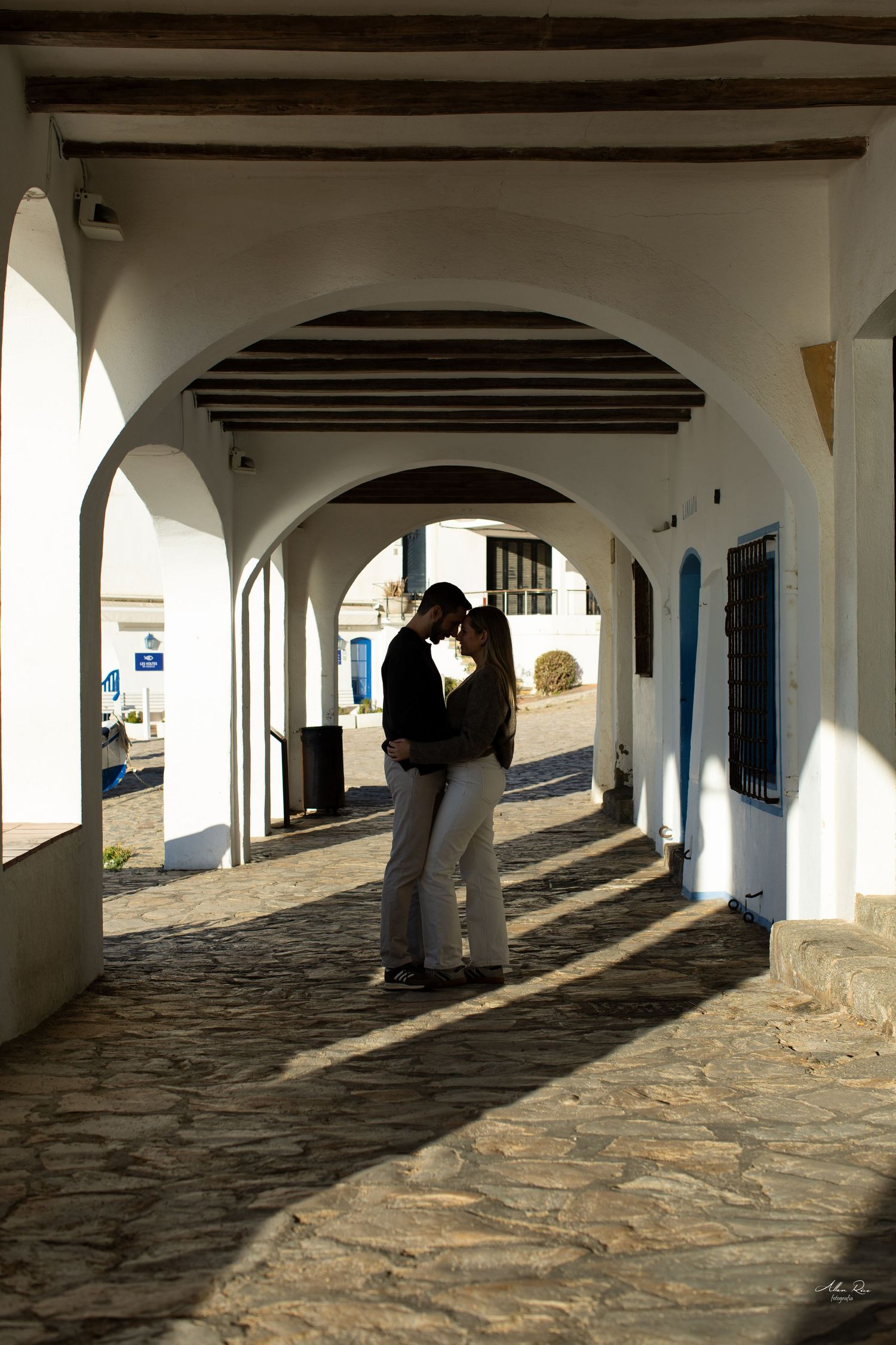 Romantic couple sharing intimate moment under white arched colonnade in charming Mediterranean village street.
