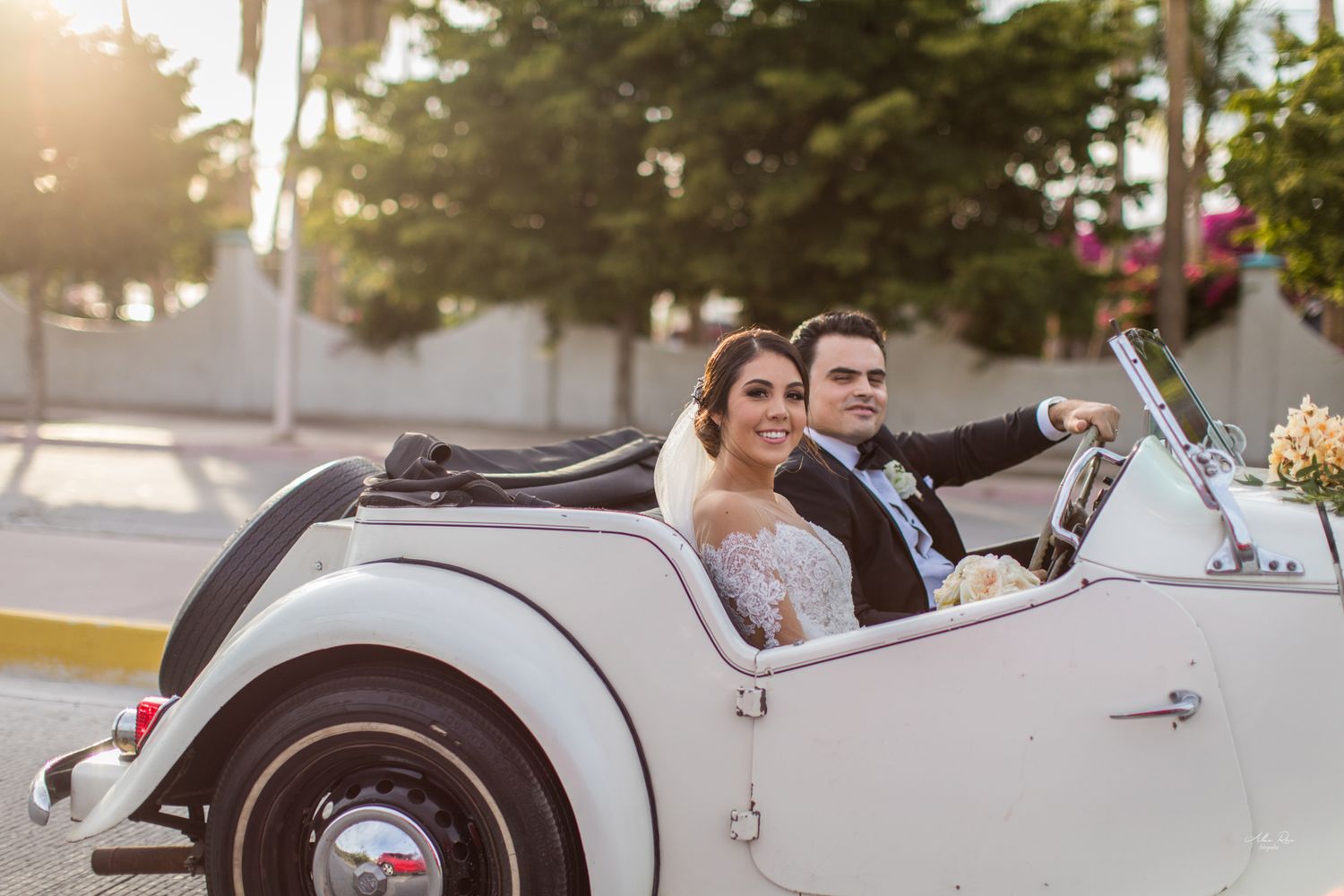 Happy bride and groom driving classic white vintage convertible car at golden hour after wedding.