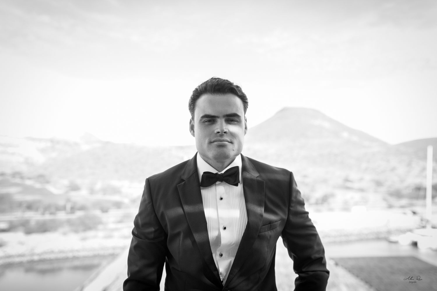 Elegant groom in black tuxedo with bow tie posing outdoors in black and white portrait.
