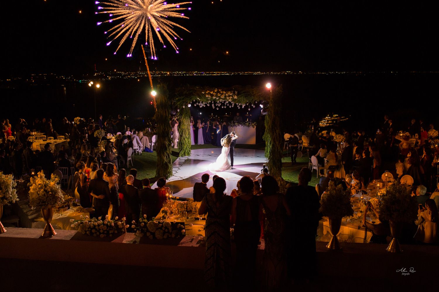 Aerial view of elegant outdoor wedding reception with fireworks display illuminating the night sky above dancing couple.