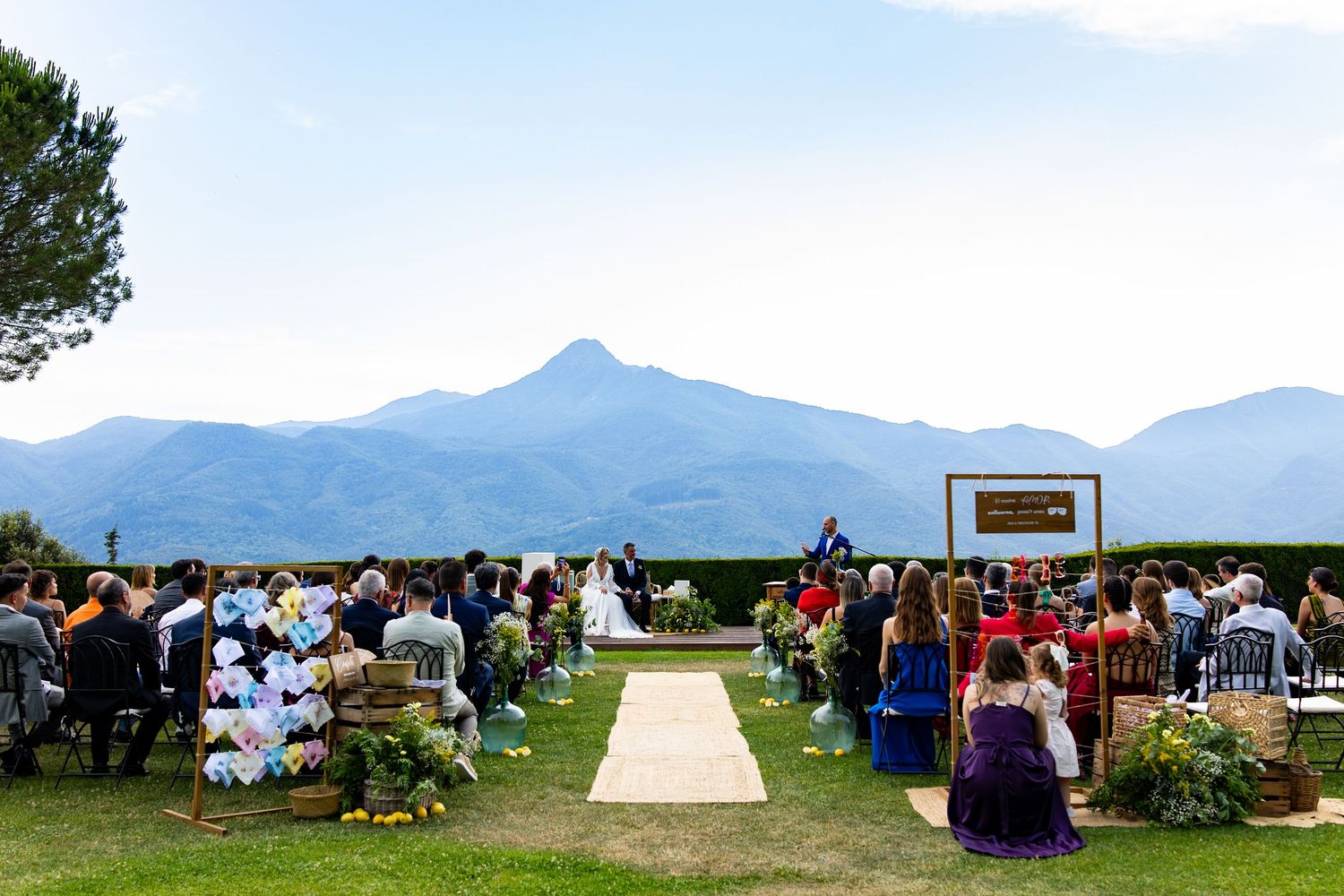 Outdoor wedding ceremony with guests seated on lawn, mountain backdrop and blue sky.
