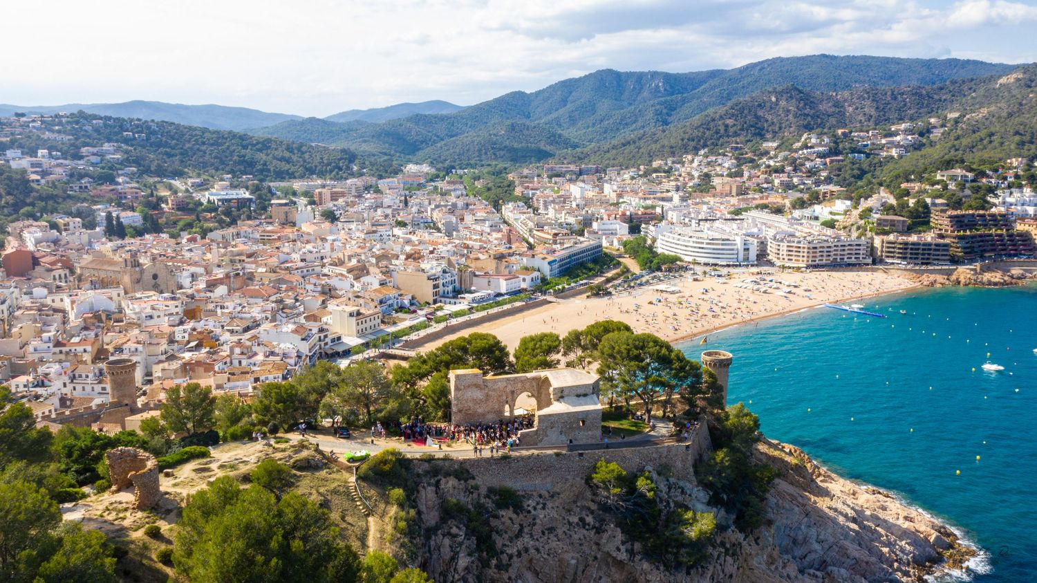 Aerial panoramic view of a picturesque Mediterranean coastal town with castle ruins and beach.