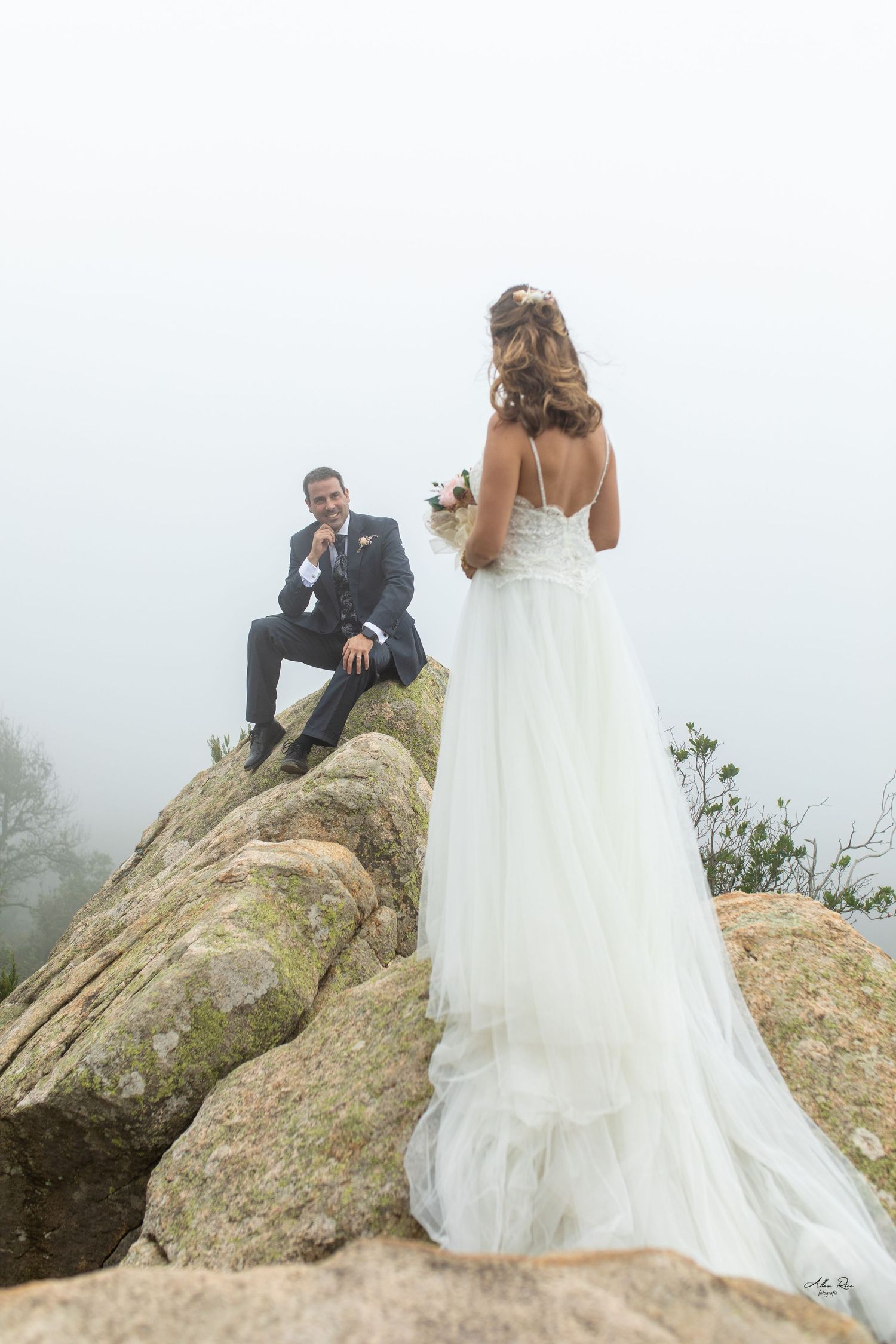 Groom sits on rocky hilltop smiling at bride in misty outdoor setting.