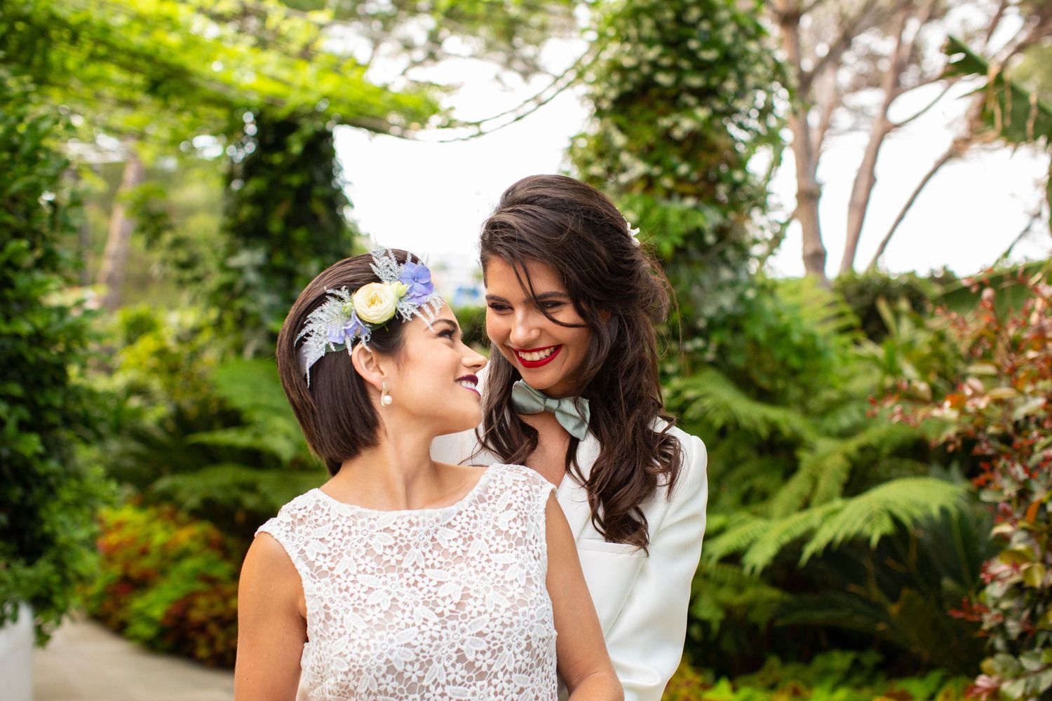 Two brides smile at each other in a lush garden, one in a lace dress with floral headpiece and one in a white blazer.