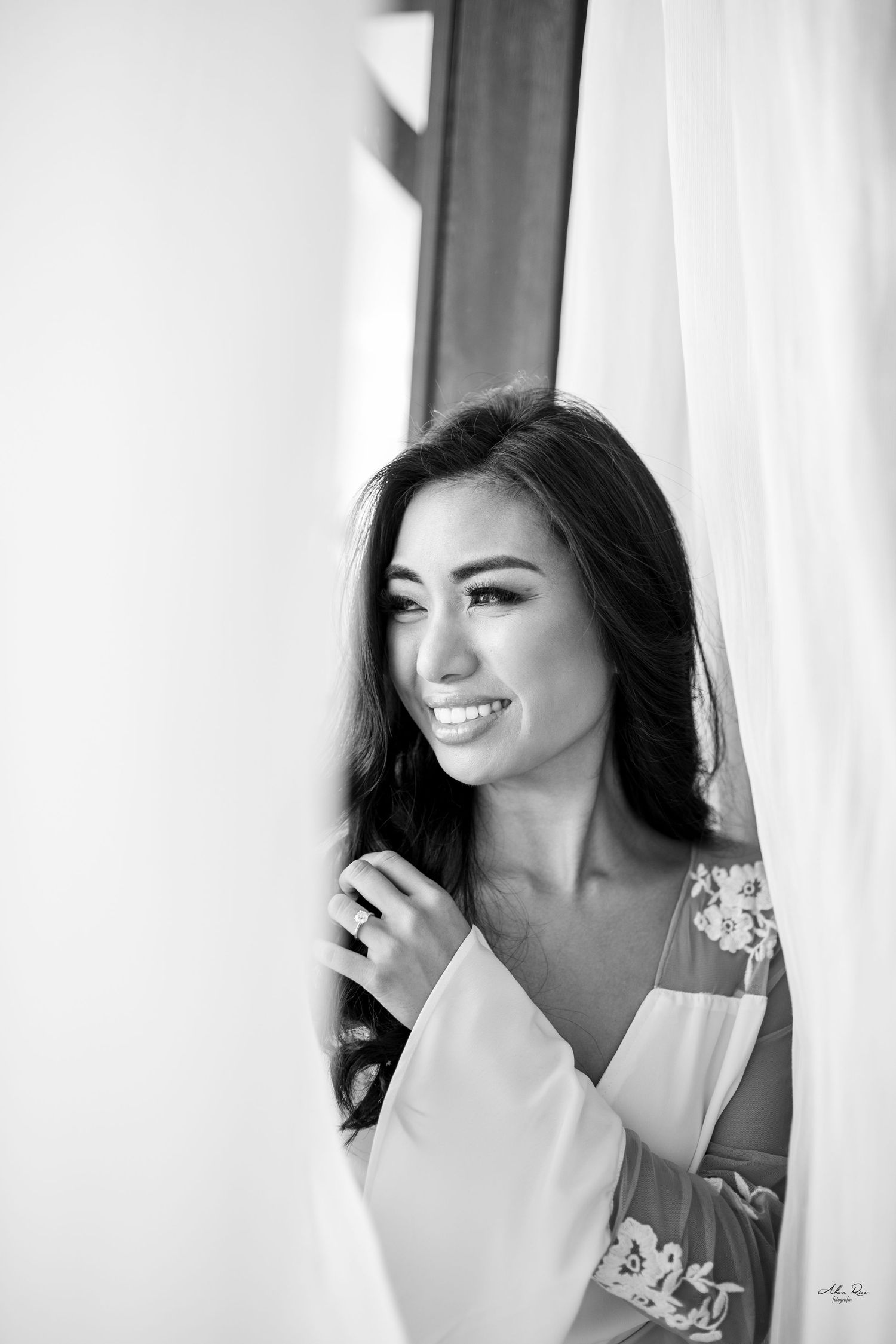 Beautiful smiling bride in floral robe posing by curtains in black and white portrait.