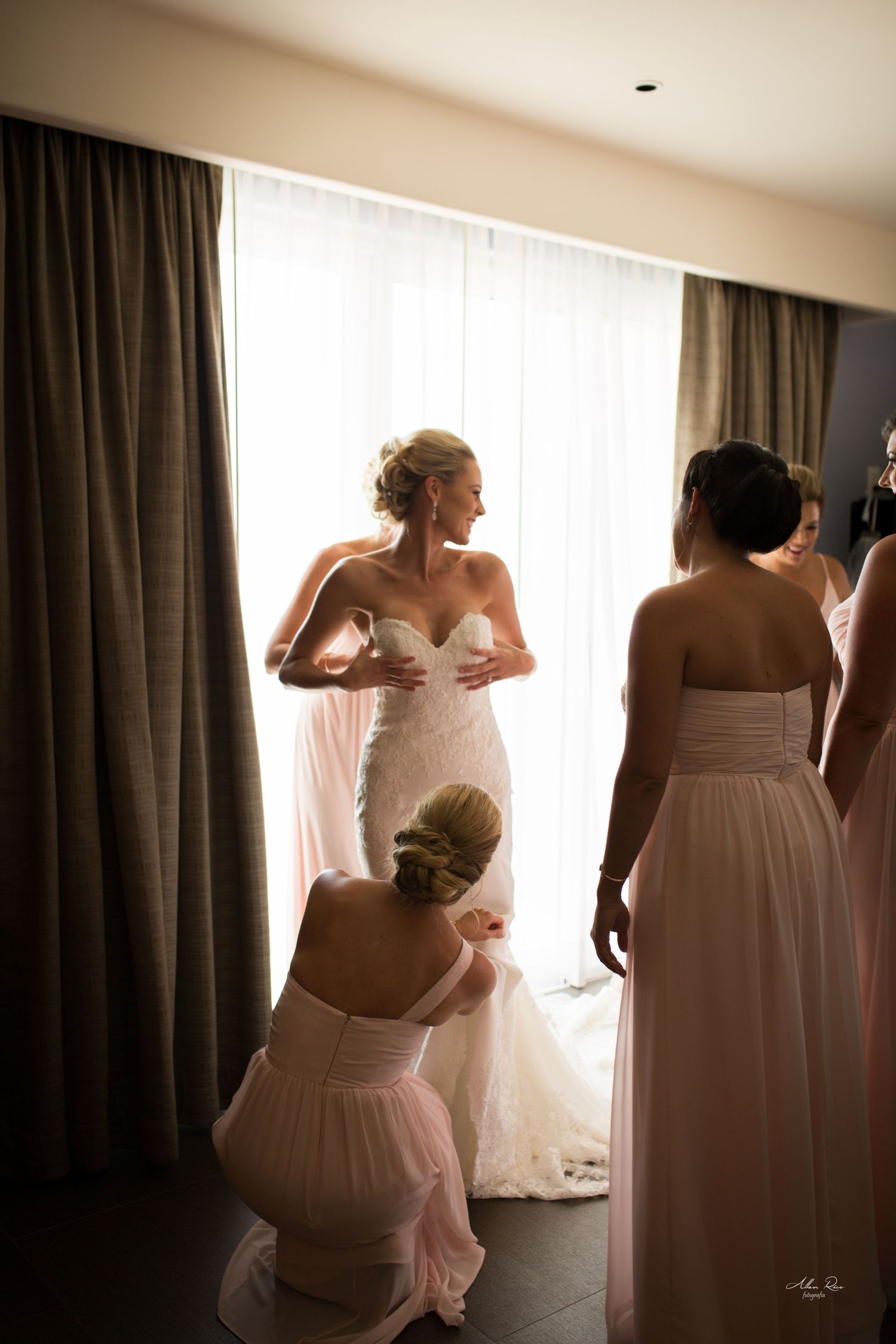 Bridesmaids helping bride into white lace wedding gown in hotel room before ceremony.
