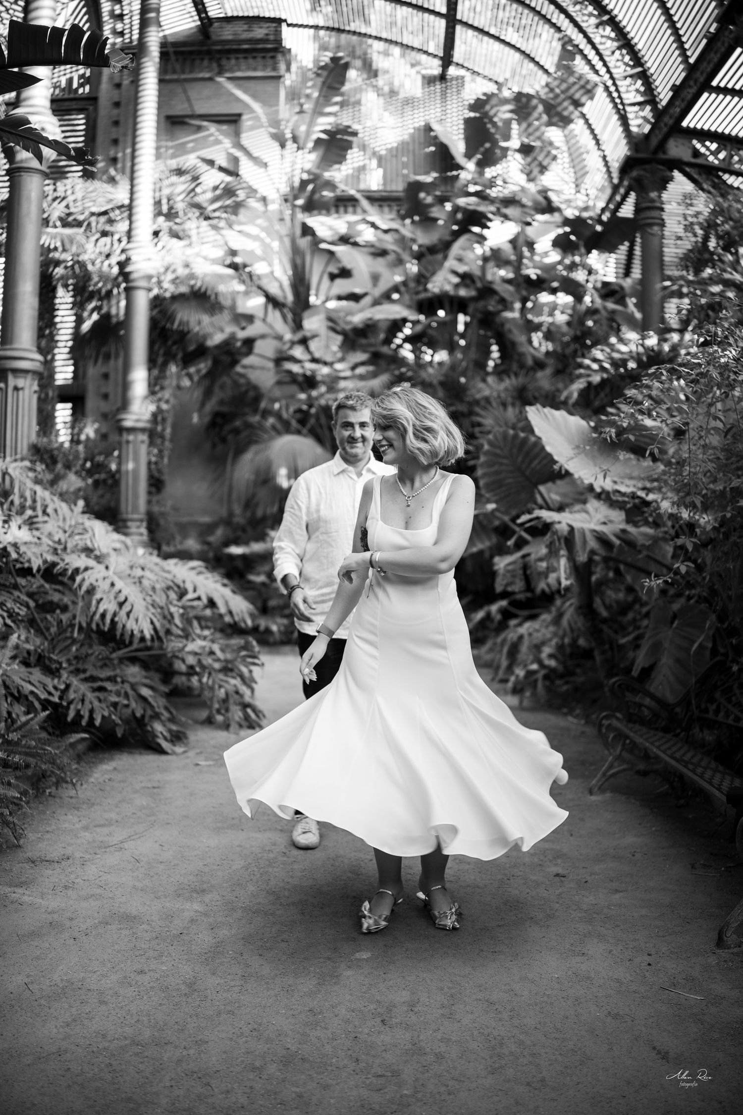 Black and white photo of a joyful couple dancing in a grand greenhouse with lush tropical plants.