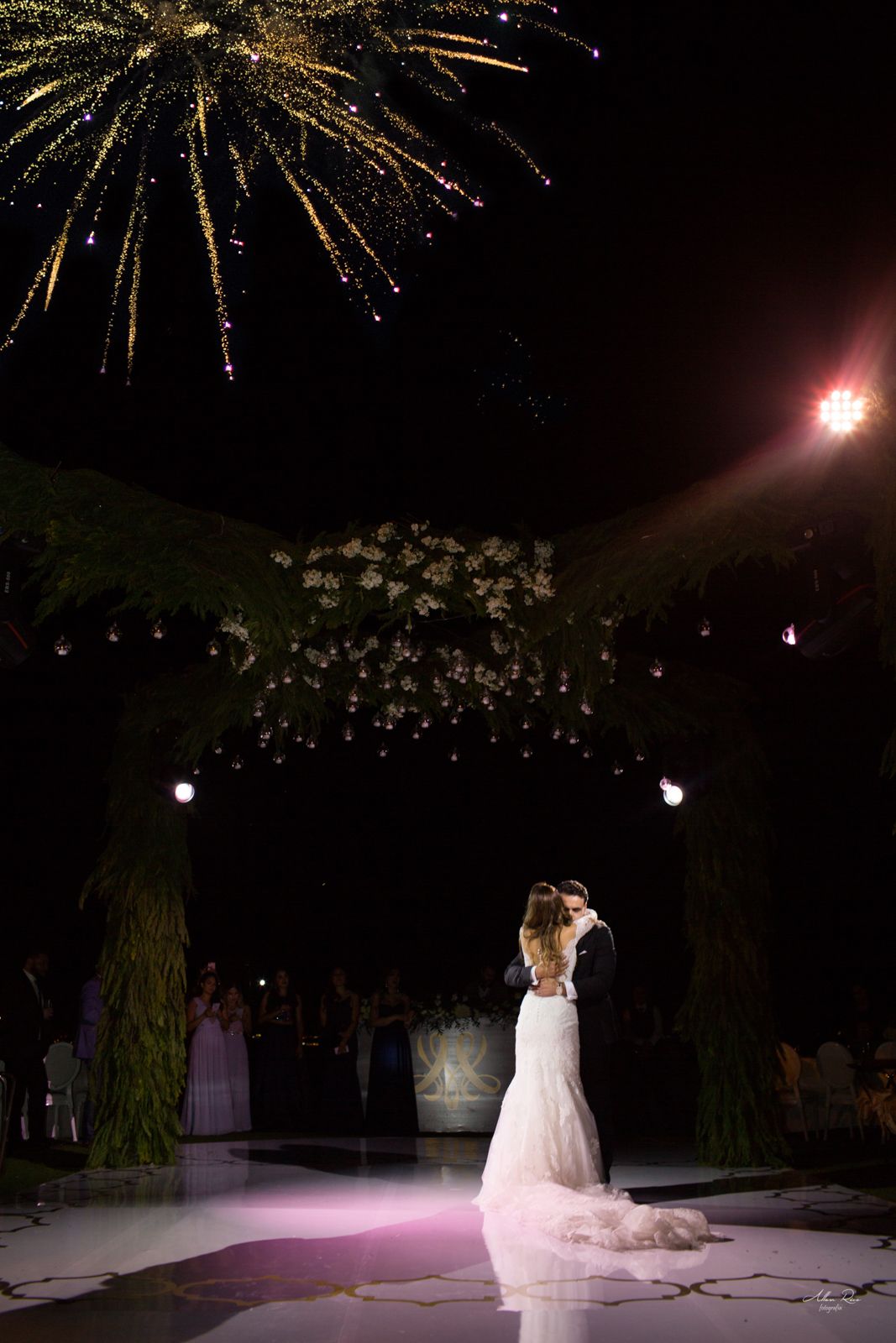 Bride and groom share first dance under fireworks and floral arch at night.