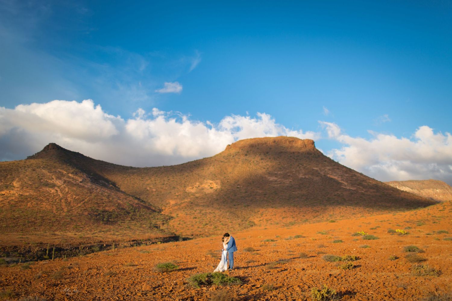 Wedding couple embraces in vast orange desert landscape beneath dramatic volcanic mountain and blue cloudy sky.
