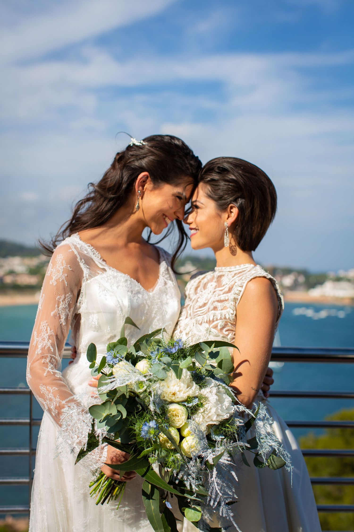 Two brides in white wedding dresses share an intimate moment touching foreheads while holding a bouquet of white roses and greenery.