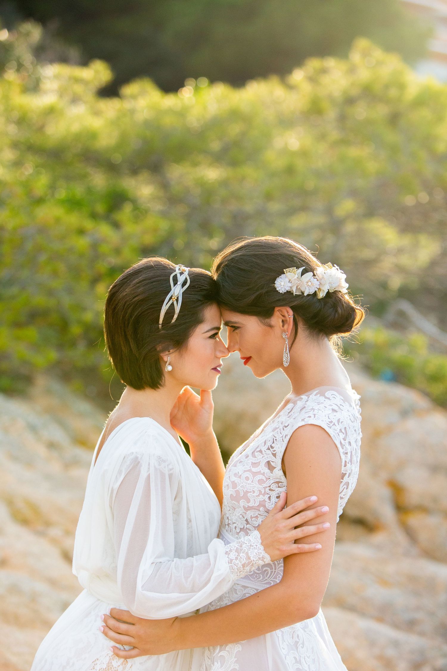 Two brides in white lace wedding dresses share a tender nose-to-nose moment in golden sunlight.