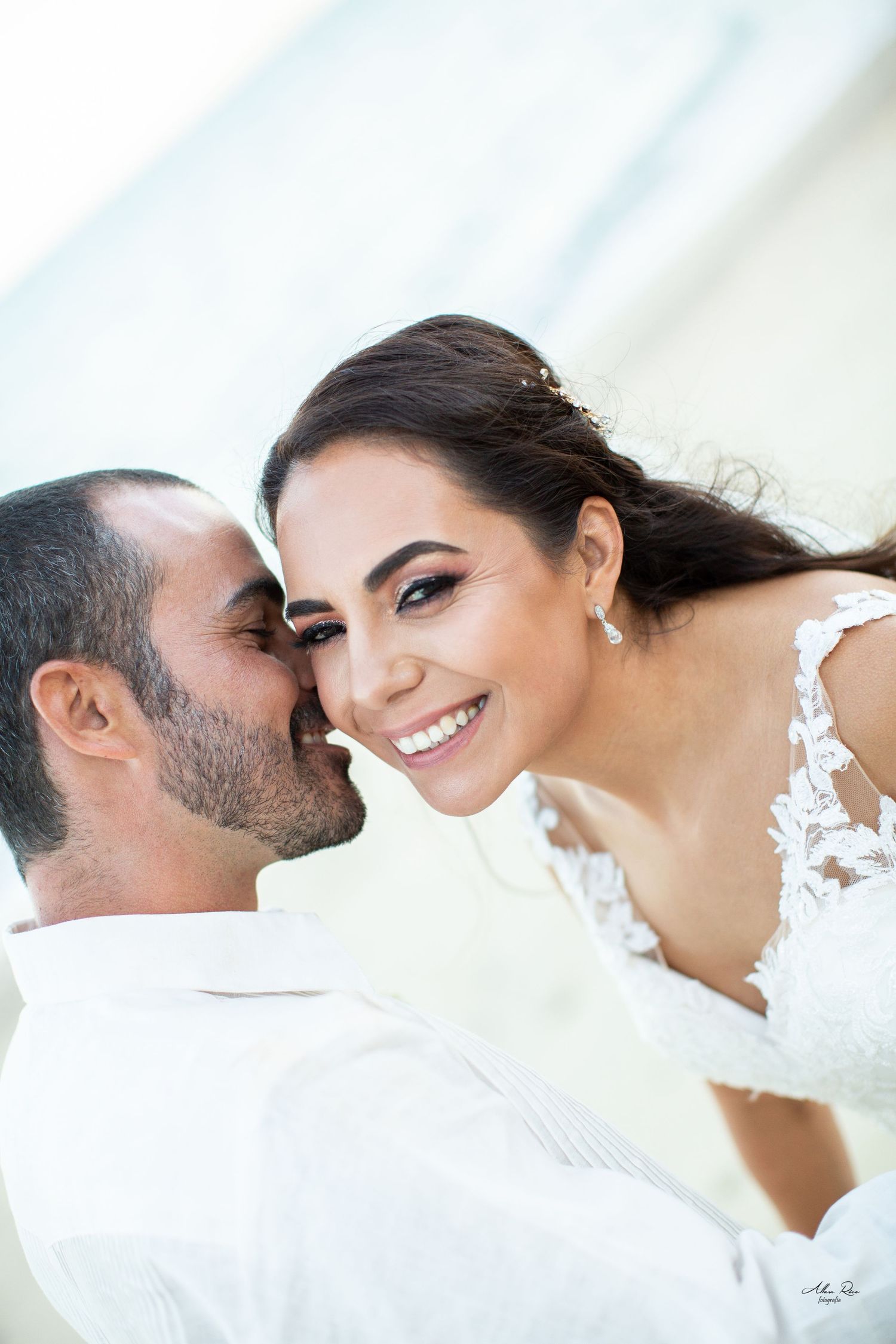 Joyful bride in lace dress smiling as groom kisses her cheek in bright outdoor wedding portrait.
