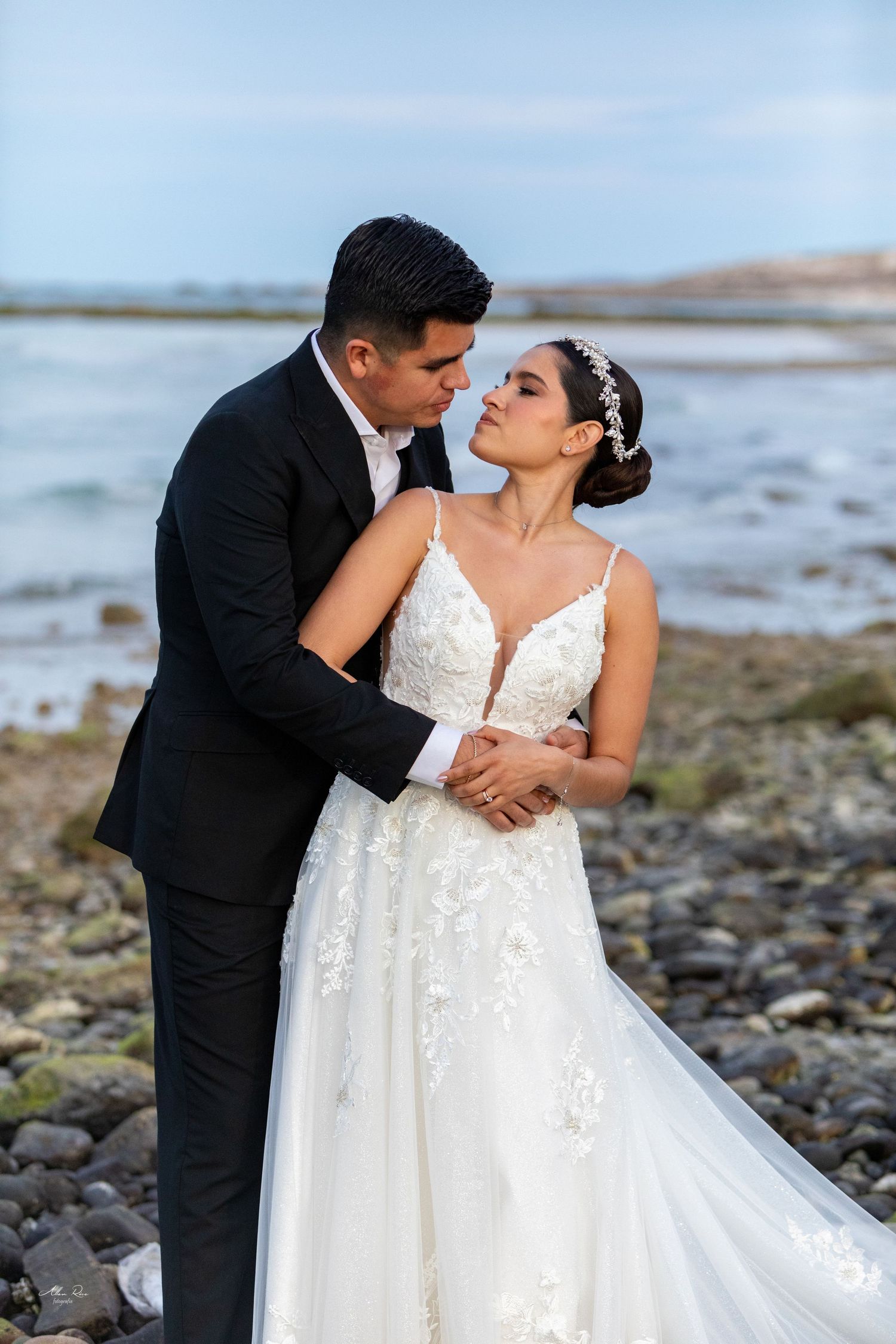 Bride and groom embrace on rocky beach shoreline, groom in black tuxedo kissing bride wearing floral lace wedding gown.
