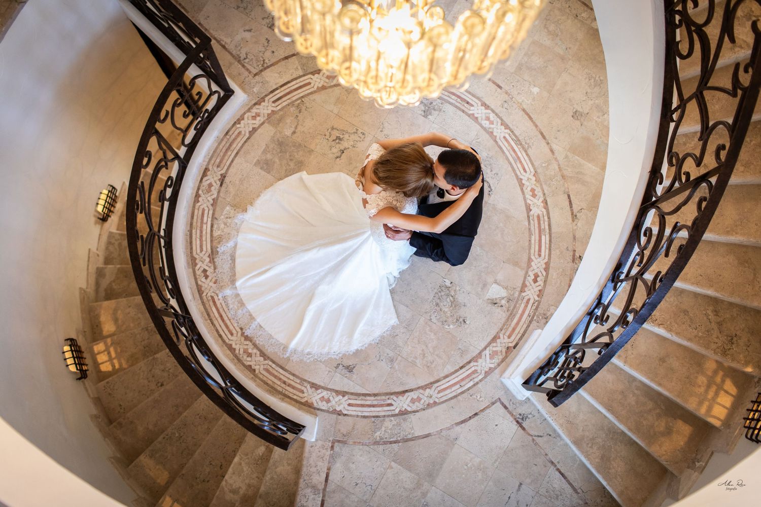 Aerial view of bride and groom embracing on grand spiral marble staircase beneath a sparkling chandelier.