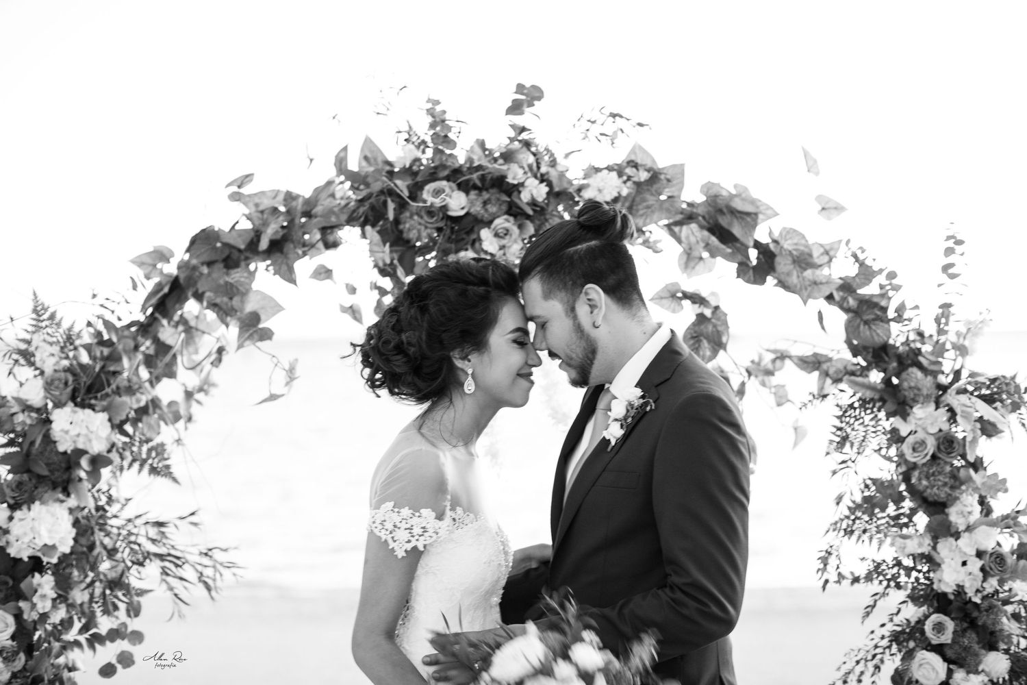 Bride and groom touch foreheads romantically under a floral arch adorned with eucalyptus and blooms.