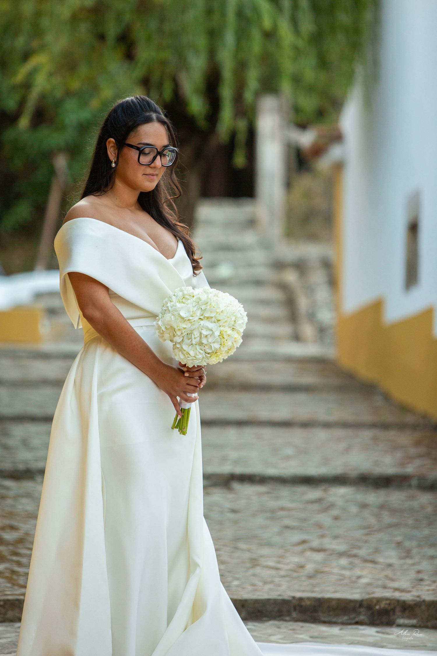 Elegant bride in off-shoulder gown holds white bouquet on stone stairway.