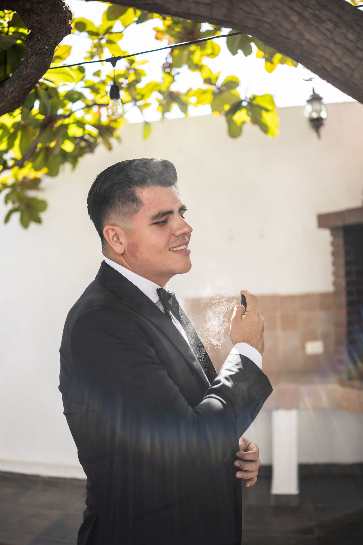 Smiling groom in black tuxedo applies cologne outdoors under a sunlit tree with string lights.