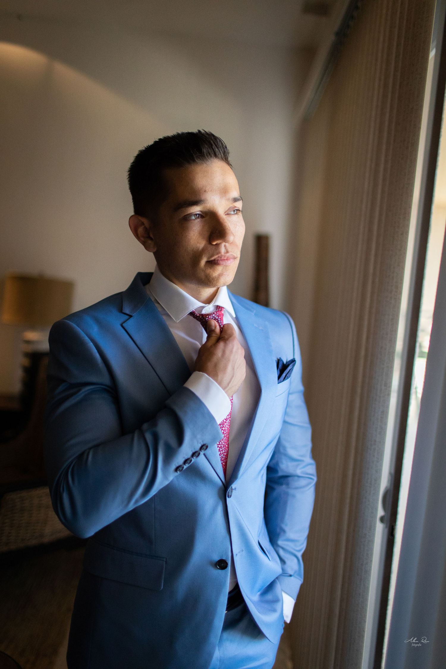 Handsome groom in light blue suit adjusting pink tie while standing by hotel window.