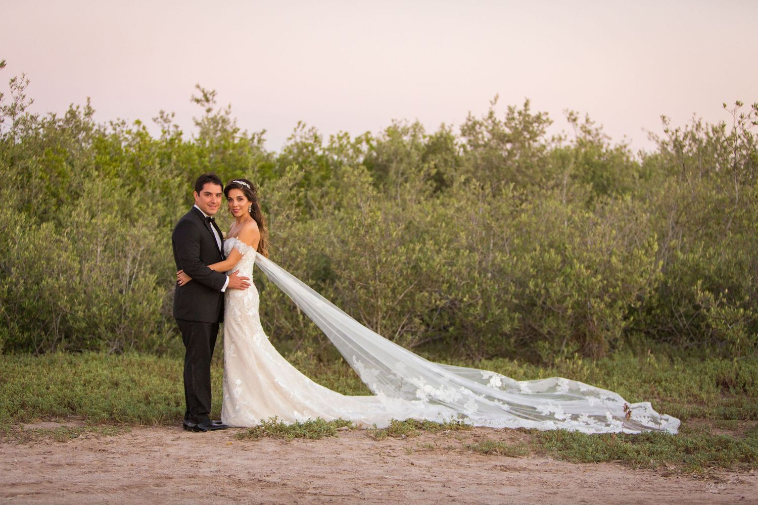 Bride and groom posing in nature with long bridal veil trailing on sandy path at sunset.