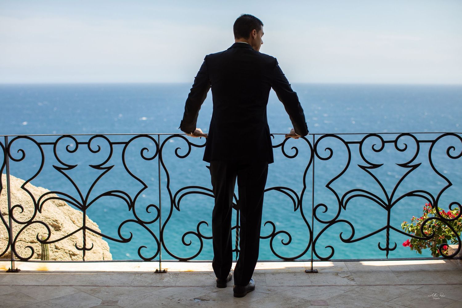 Groom in black suit stands alone at ornate wrought iron balcony overlooking turquoise ocean and clear sky.