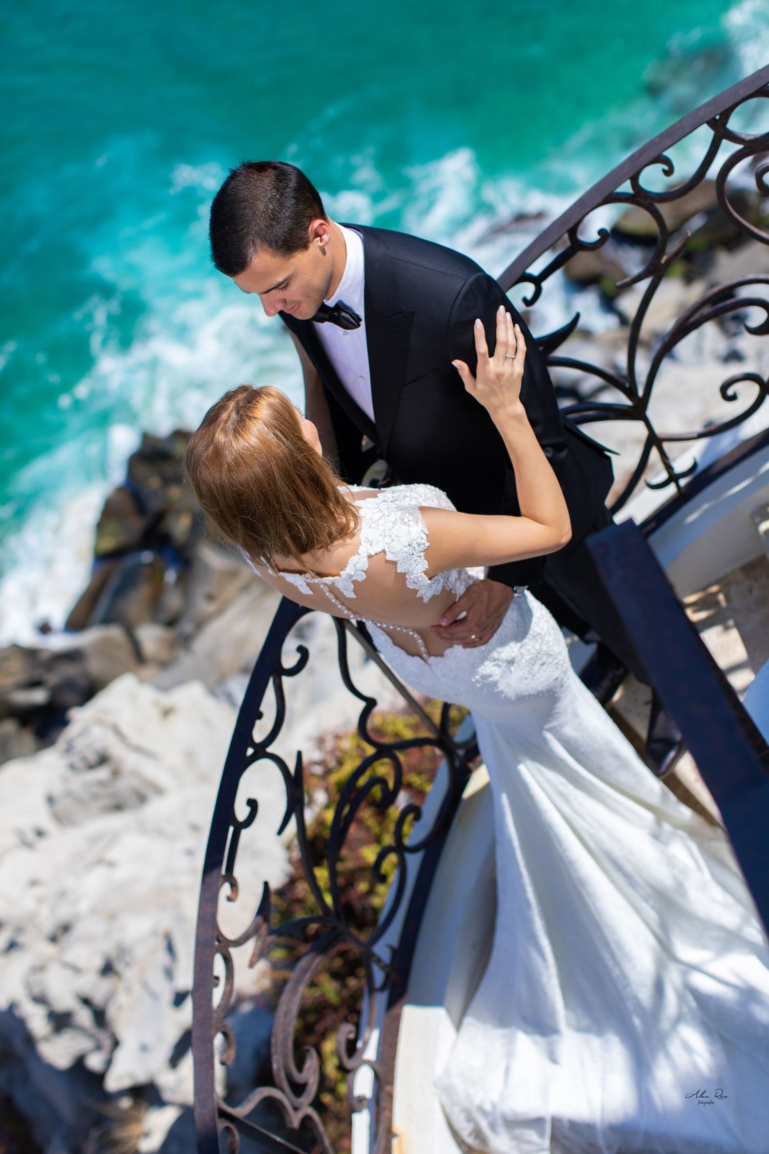 Aerial view of bride and groom sharing intimate moment on ornate iron balcony above turquoise ocean waters.
