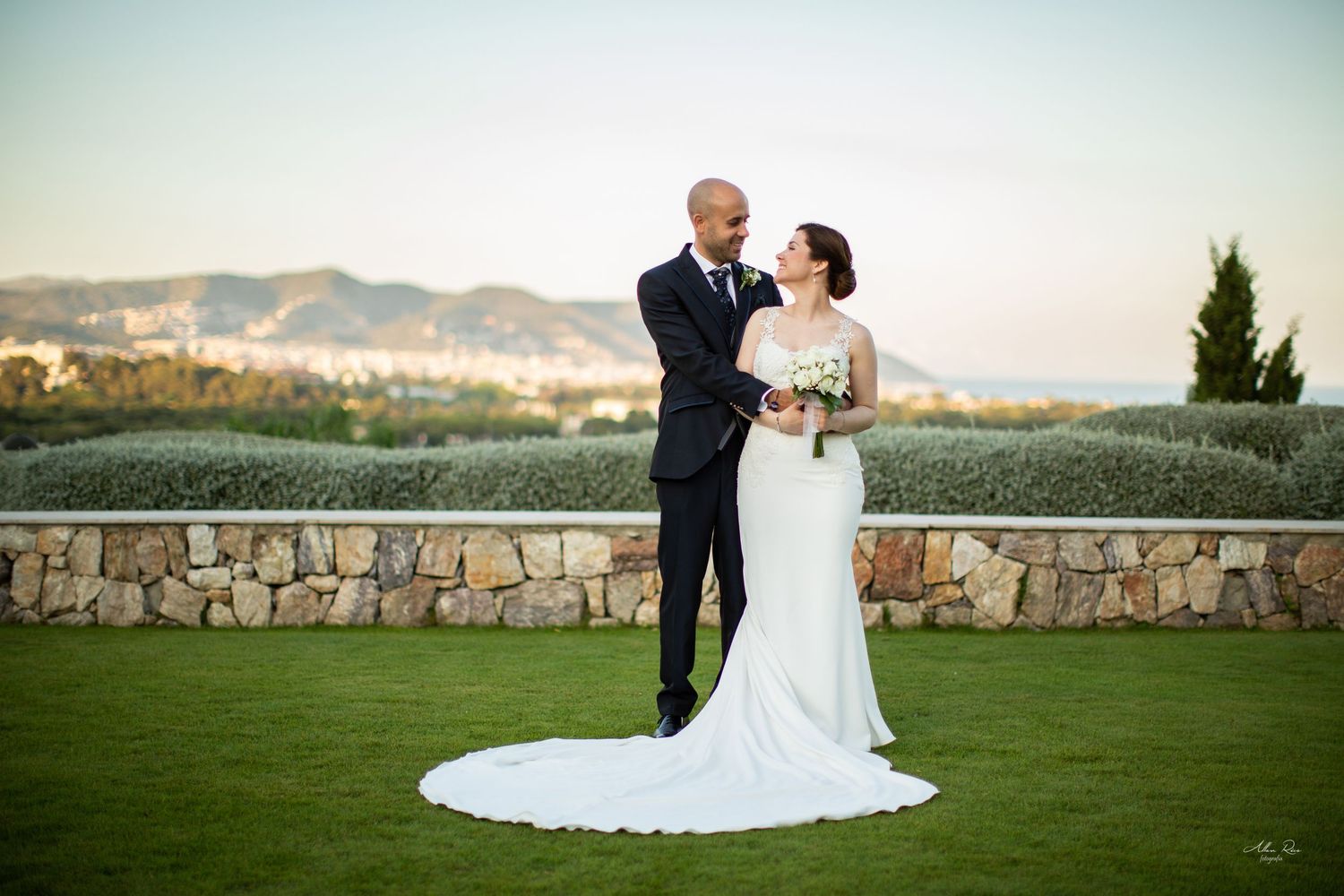 Elegant bride and groom posing on lush green lawn with scenic mountain backdrop at sunset.