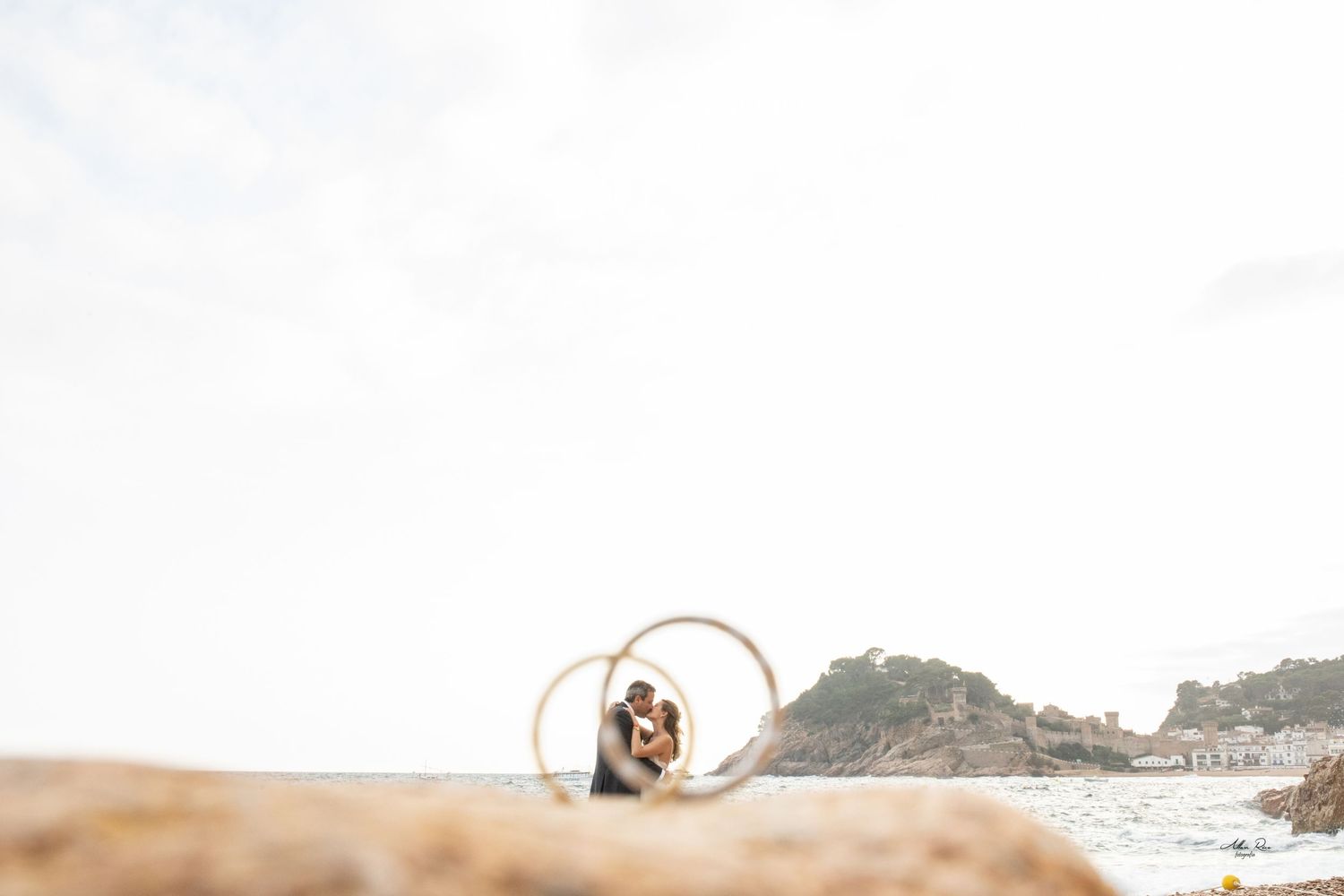Couple embraces framed by golden rings on a rocky coastal beach under bright sky.