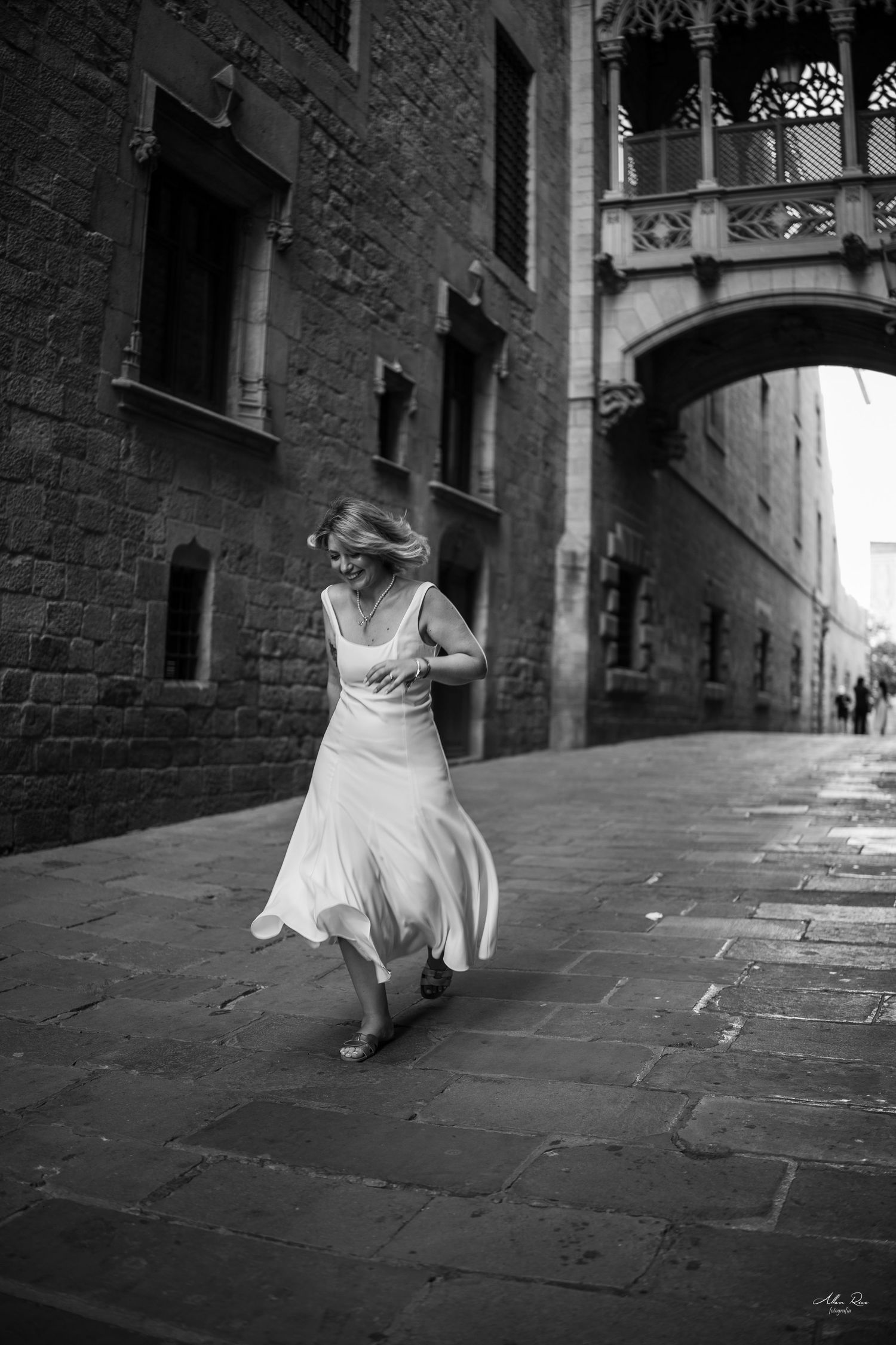 A woman in a flowing white dress twirls joyfully in a black-and-white photo on a historic Barcelona street.
