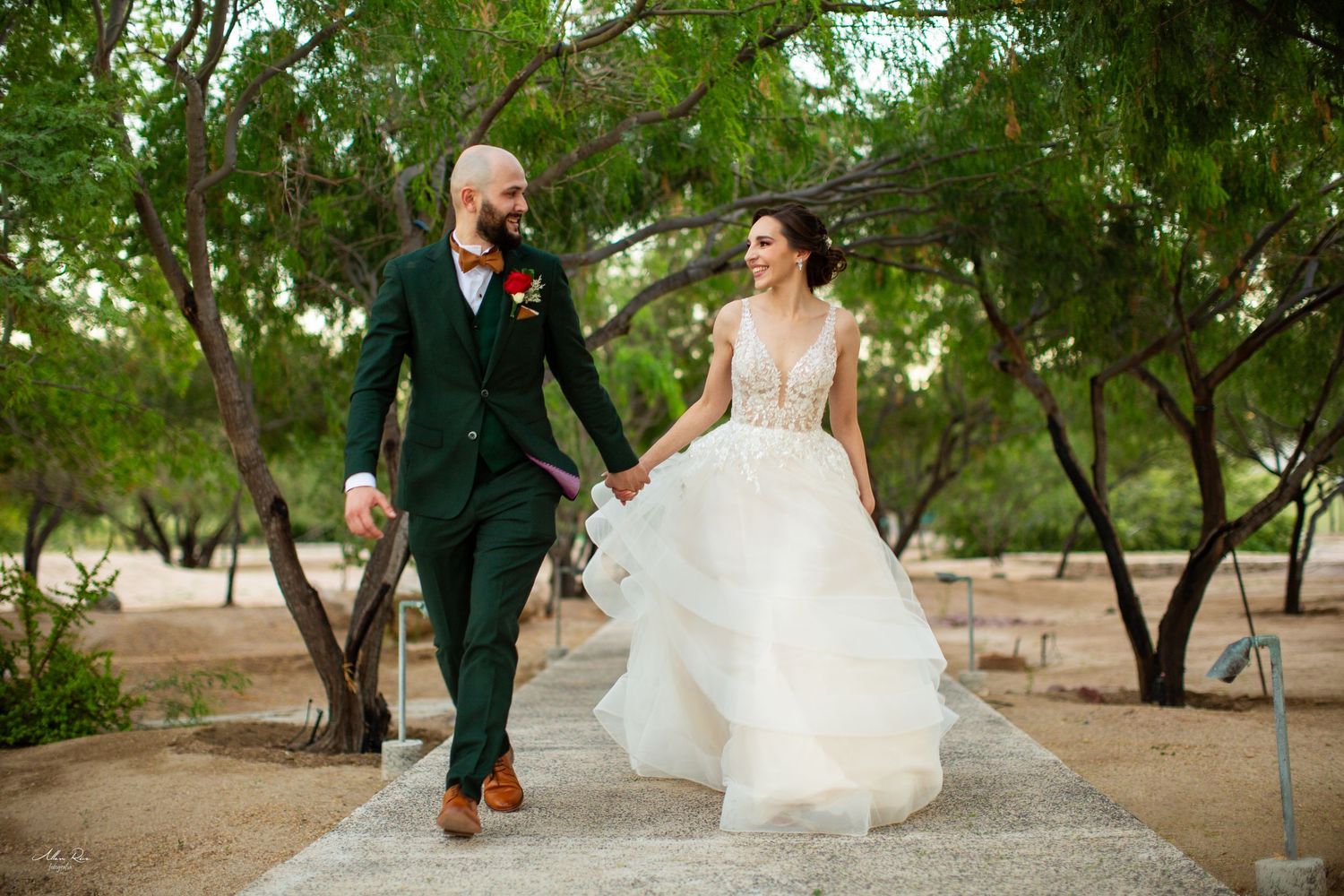 Happy bride and groom walk hand in hand through a tree-lined path on their wedding day.