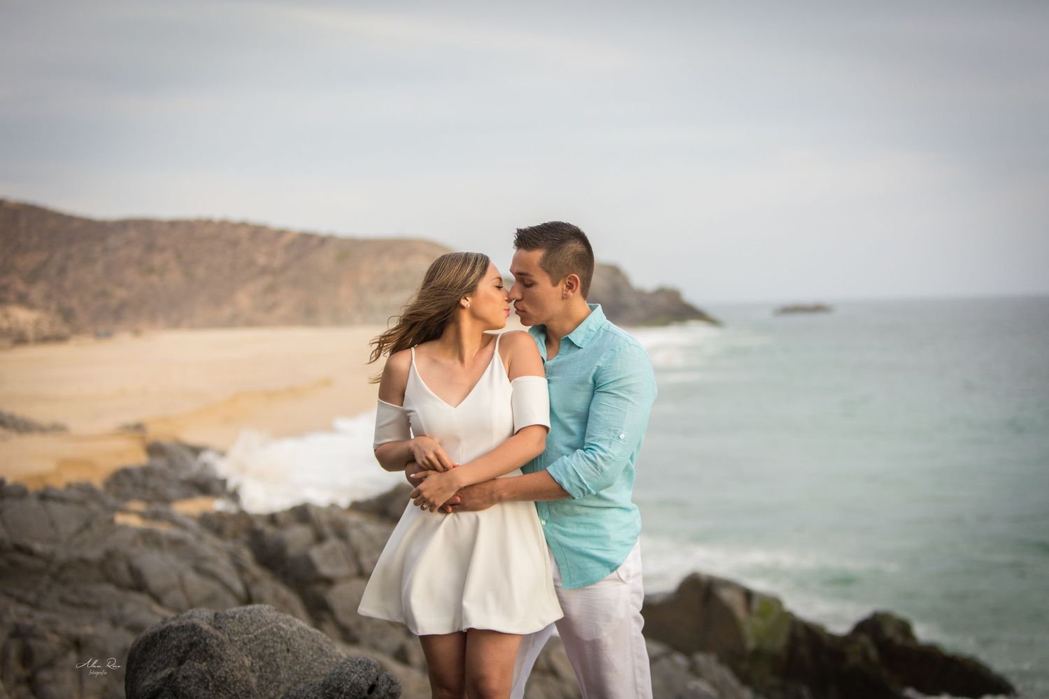 Couple shares a romantic kiss on rocky coastal cliffs with ocean waves behind them.
