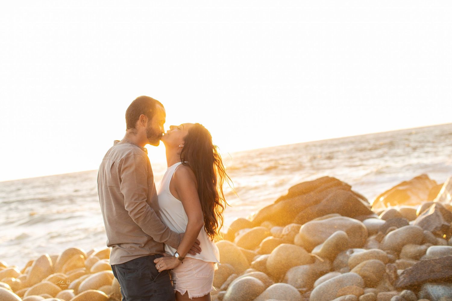 Couple sharing a kiss on a rocky beach with golden sunset light and ocean waves behind them.