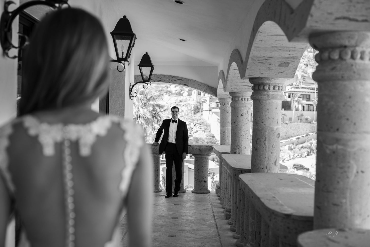 Black and white photo of groom walking toward bride through elegant arched colonnade with stone pillars.