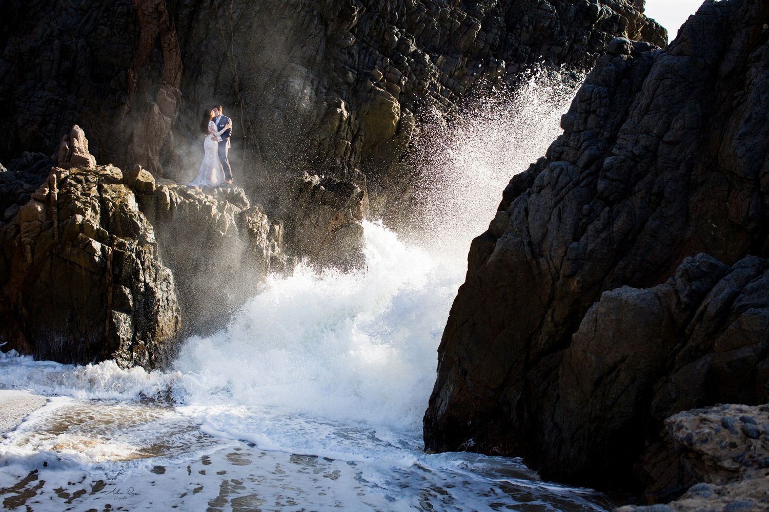 Daring wedding couple posing on rocky cliffs with dramatic crashing ocean waves below.