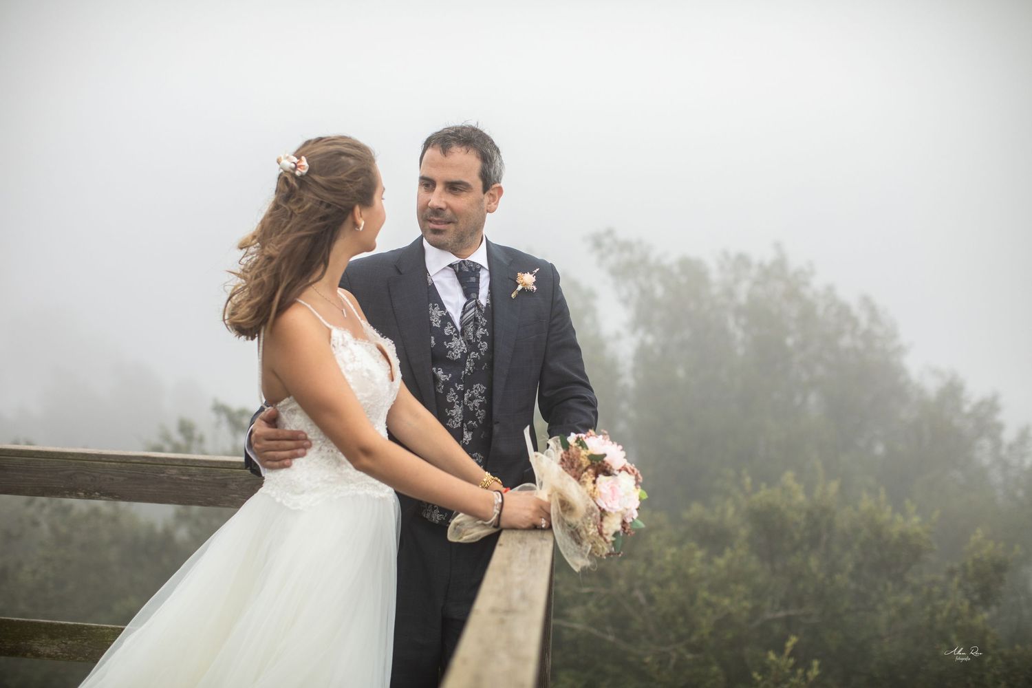 Smiling bride and groom share a tender look on foggy wooden bridge.