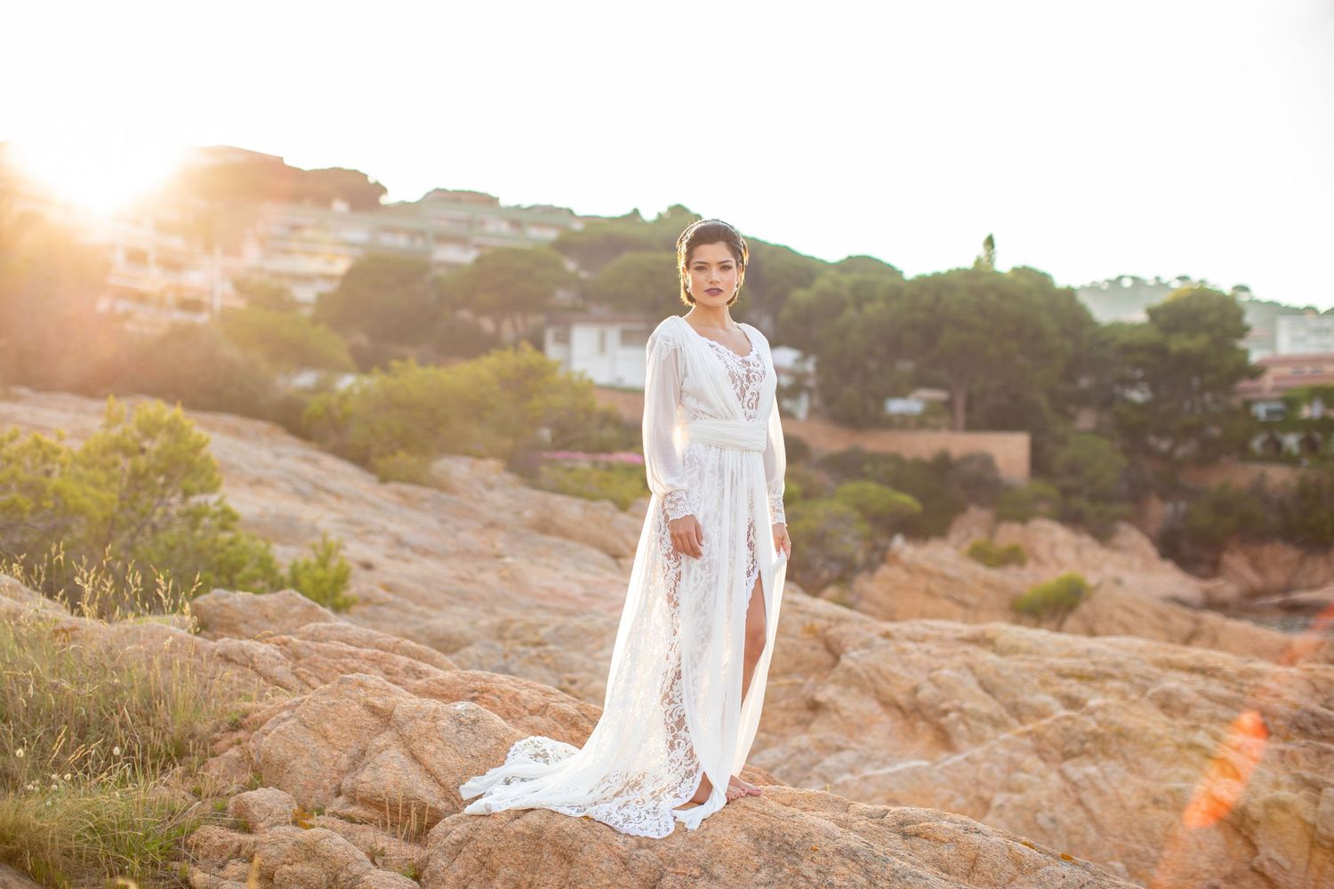 Bride in long-sleeved white lace wedding gown poses confidently on sun-drenched rocky coastal landscape.