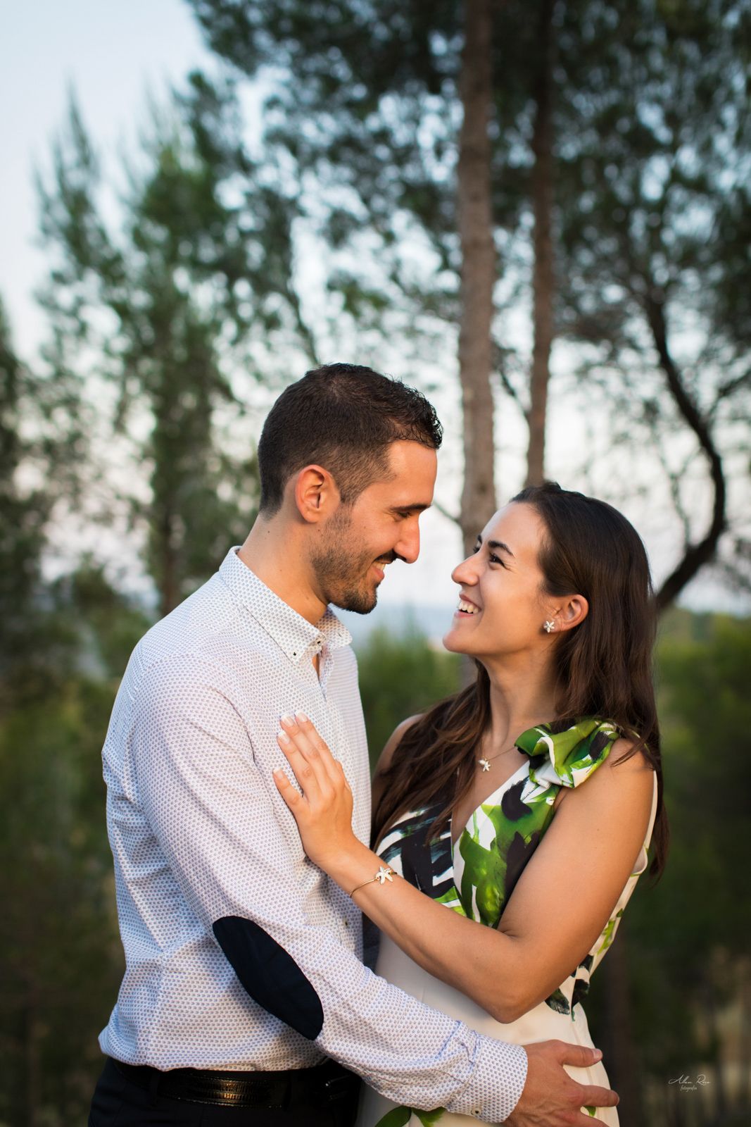 Happy couple smiles and embraces closely outdoors surrounded by green trees at dusk.