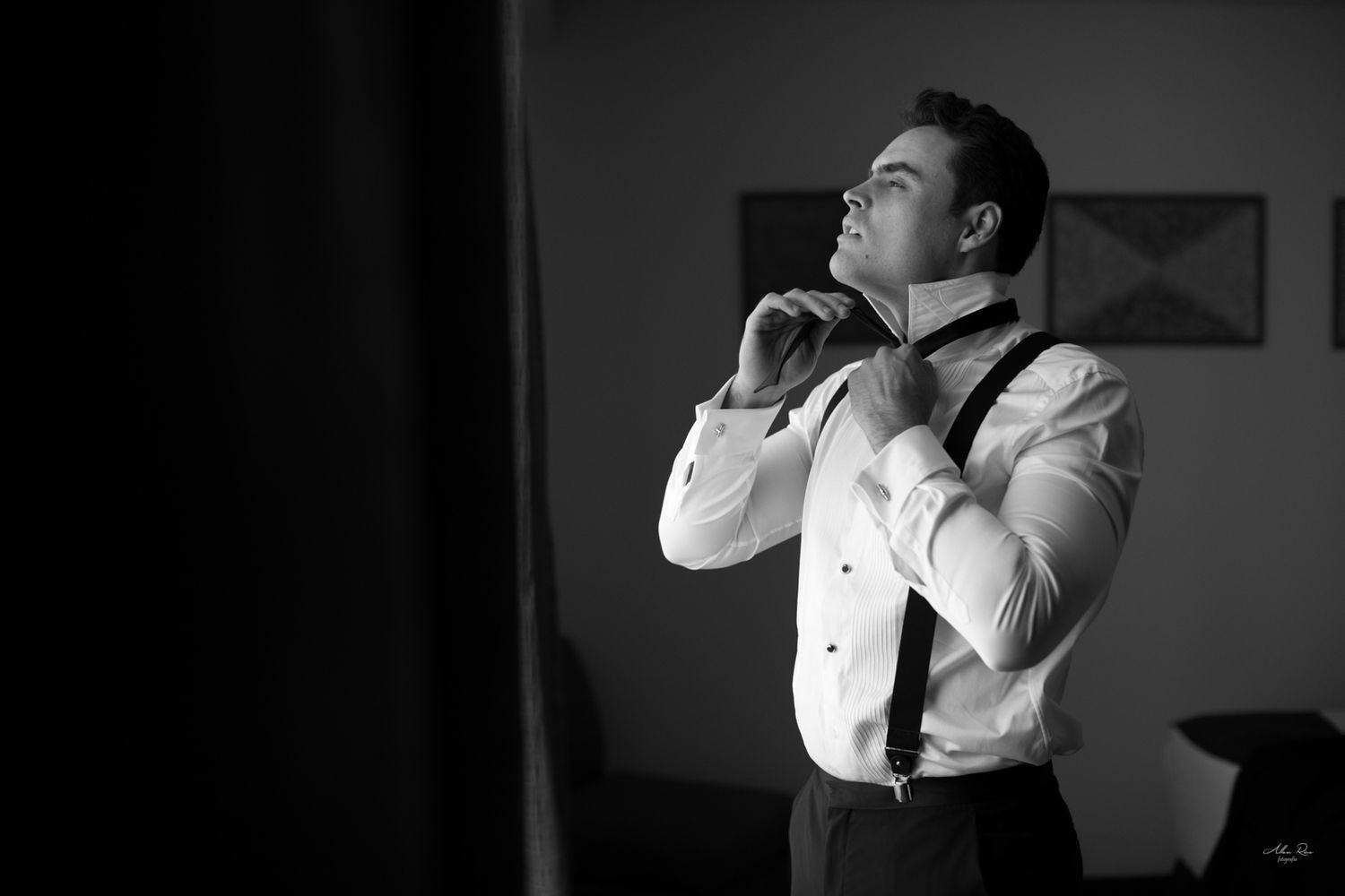 Black and white photo of groom in suspenders adjusting his bow tie while getting ready.