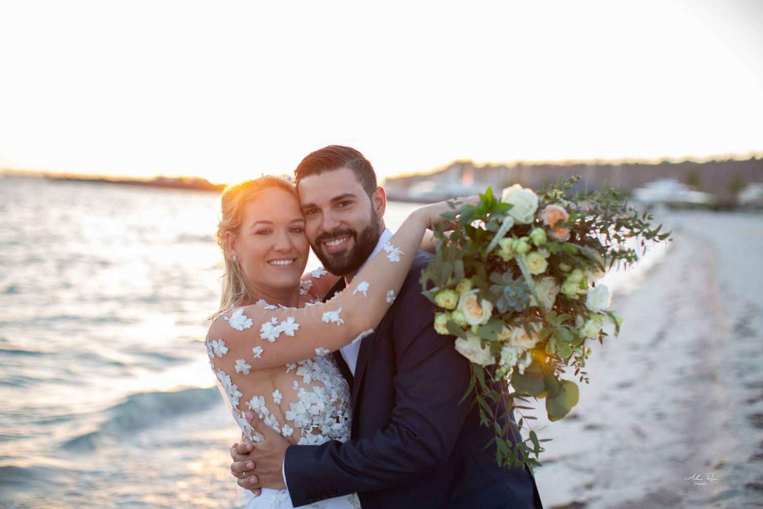 Smiling bride and groom embrace on a beach at sunset, bride holding a lush peach and green bouquet.