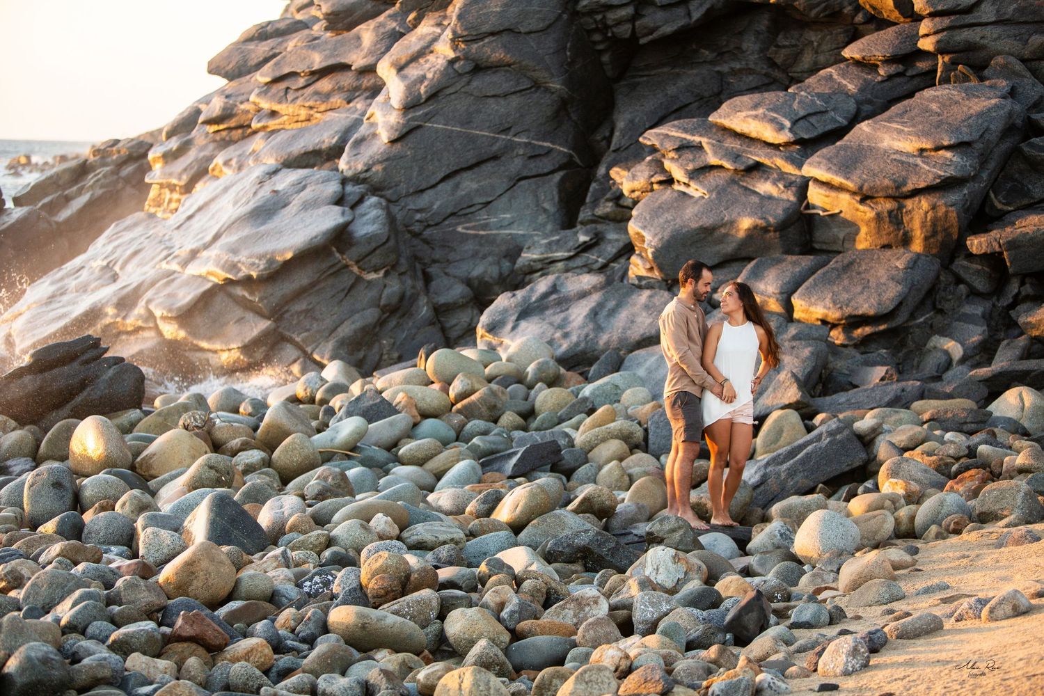 Couple standing close together on a rocky coastal beach surrounded by smooth boulders at golden hour.