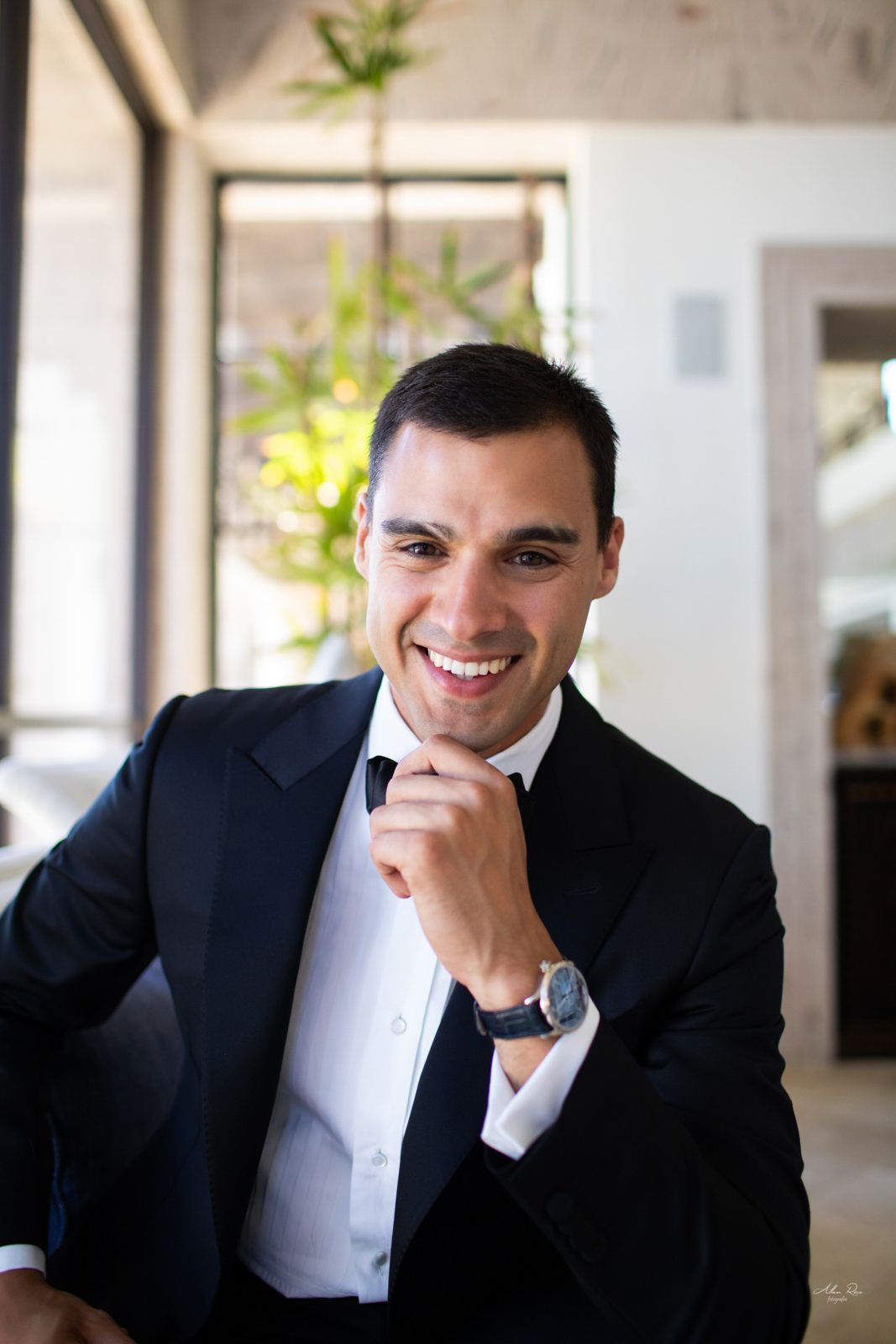 Smiling groom in black tuxedo adjusting bow tie indoors on wedding day.